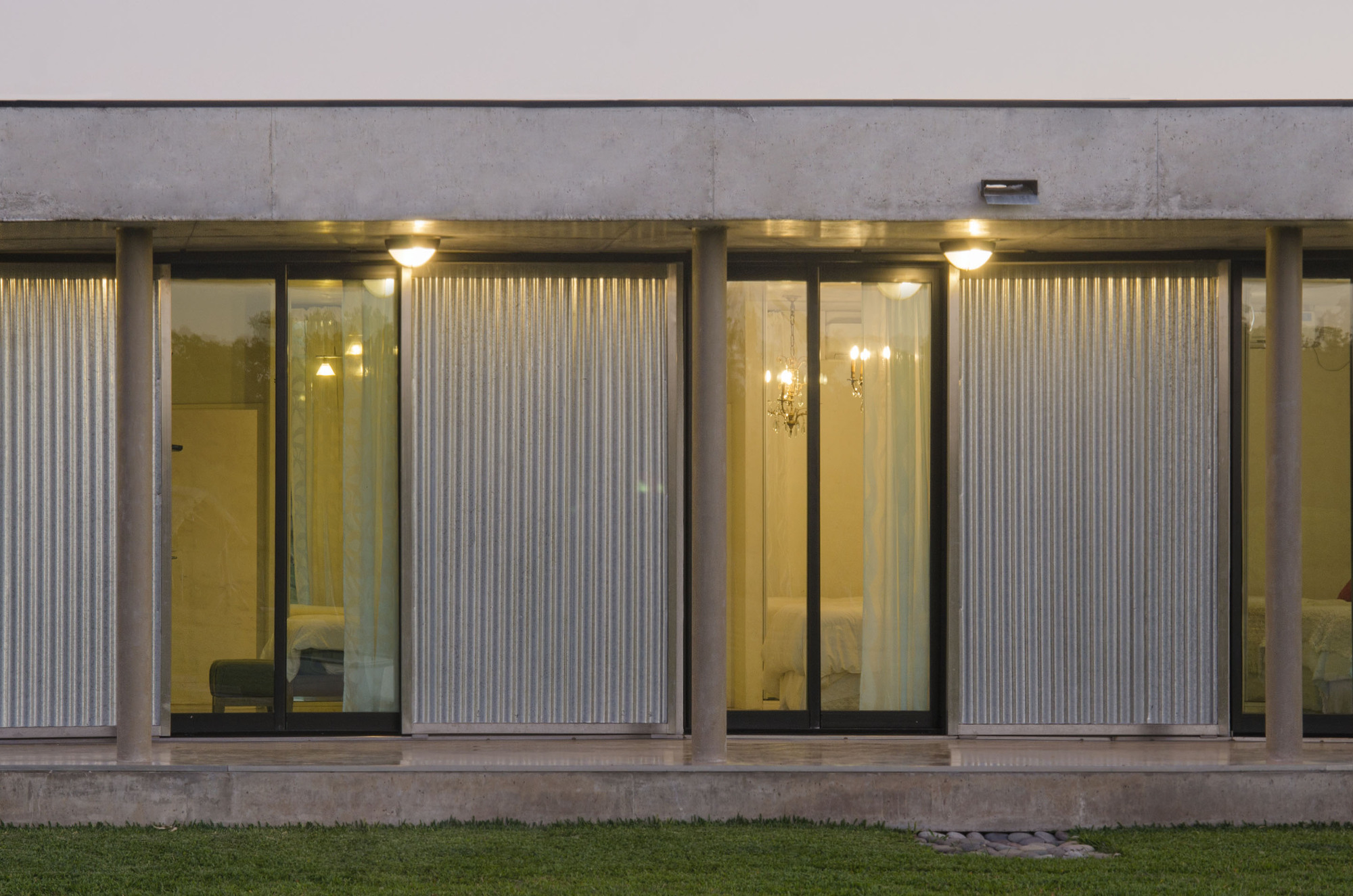 Close view of the porch at dusk, with corrugated metal panels alternating with glass doors that show warmly lit bedrooms behind