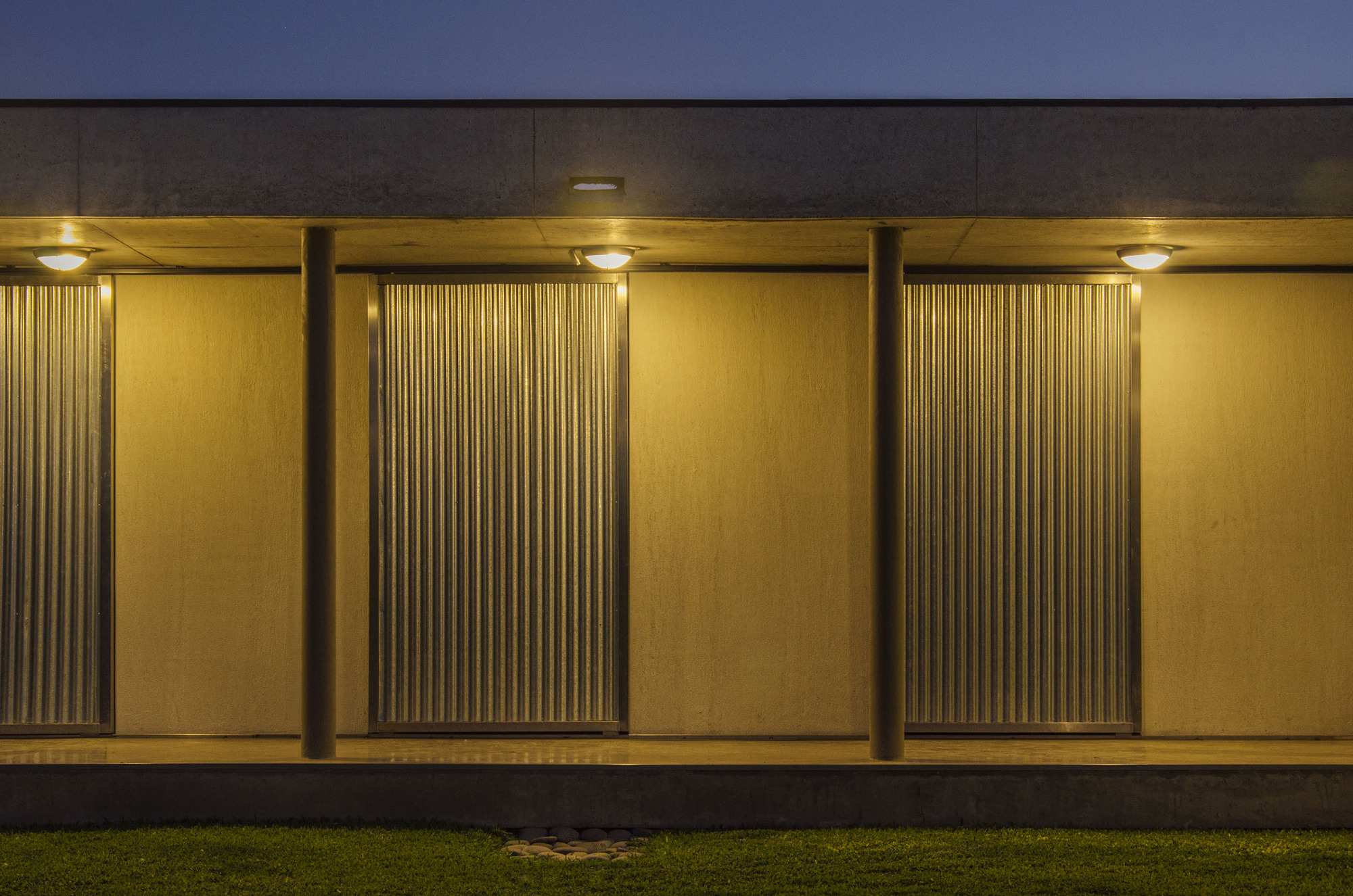 Night view of the porch wall with corrugated metal panels between round columns, lit by ceiling lamps above a lawn edge