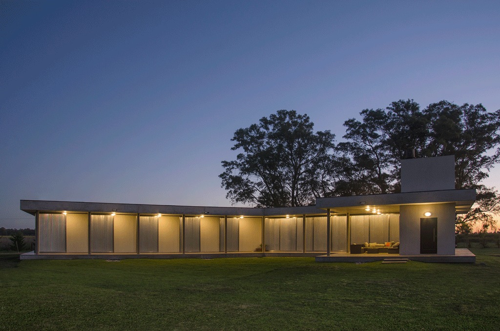 Long L-shaped house with corrugated shutters and downlights glowing under a dusk sky, with tall trees behind the lawn