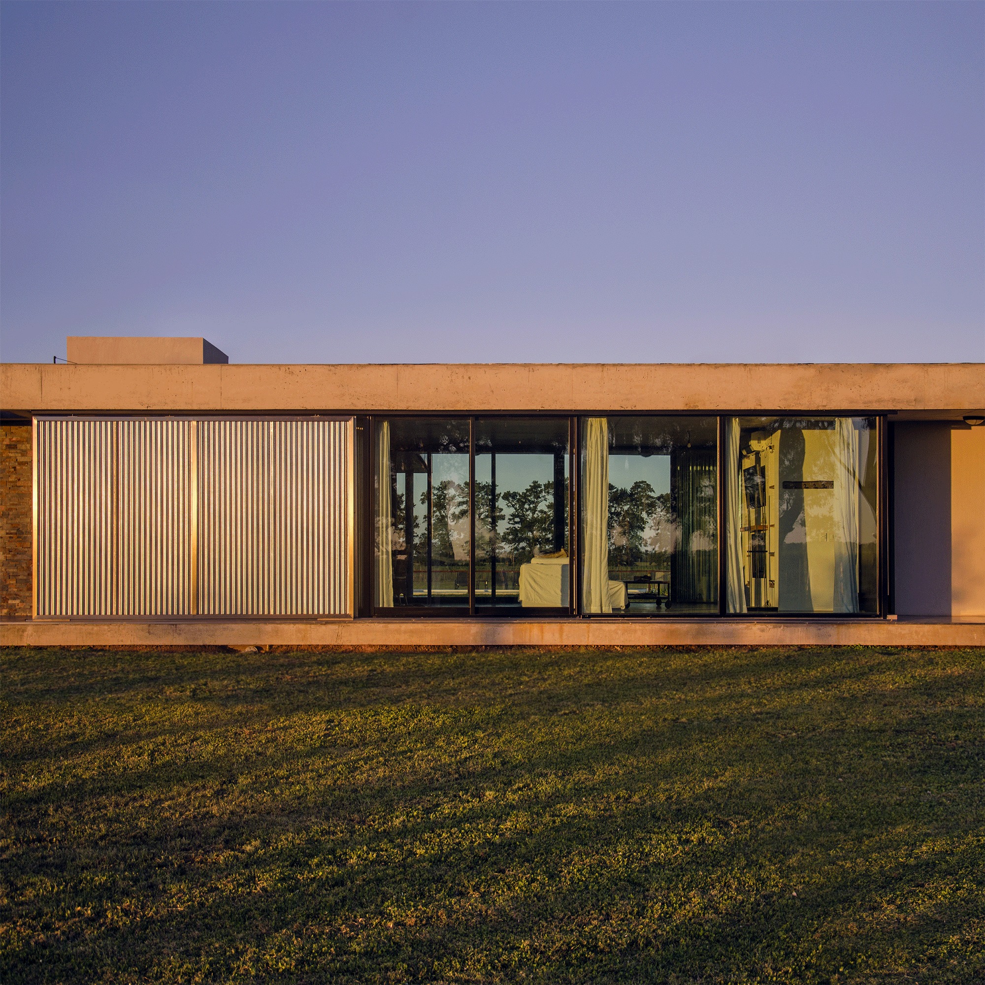 Concrete-framed facade with sliding corrugated metal shutter beside glazed doors and curtains, lit by low evening sun on the grass
