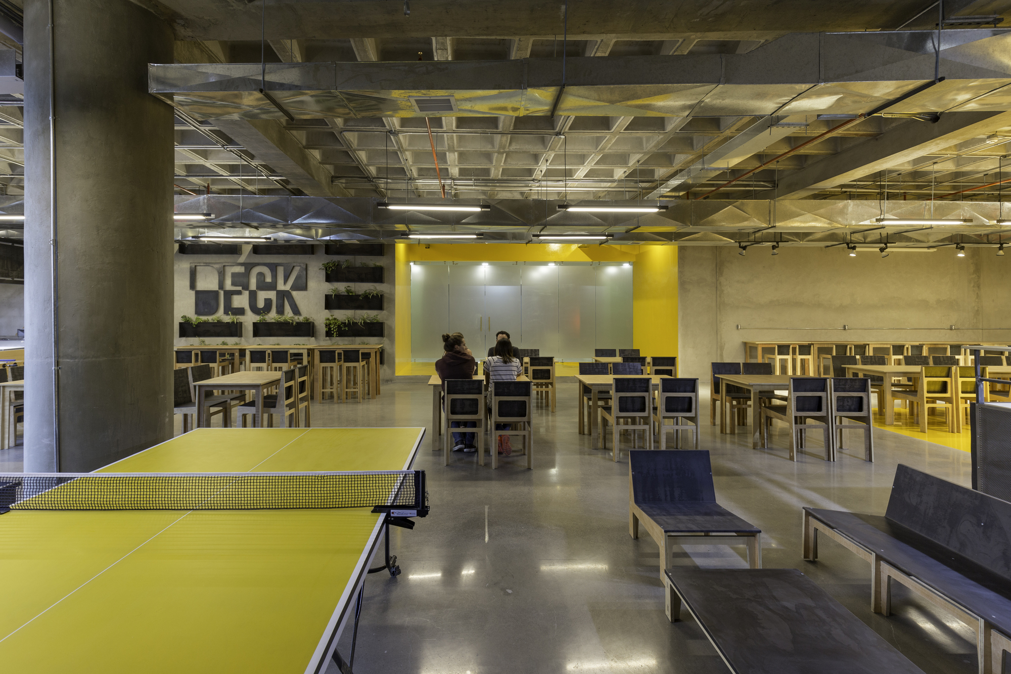 Yellow ping-pong table in the foreground of a dining hall with plywood chairs, a yellow wall and two people seated at a table