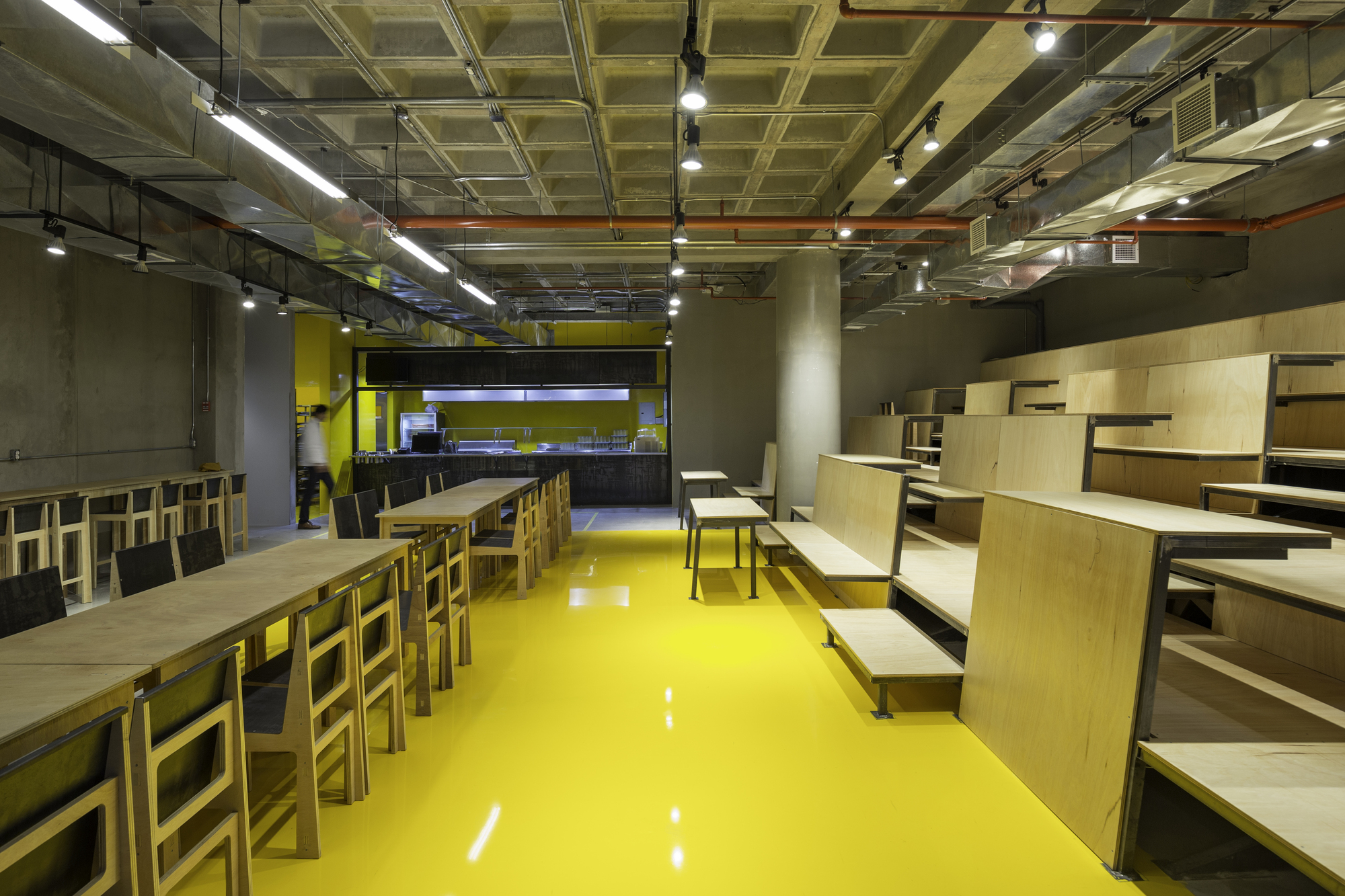 Glossy yellow floor running between plywood tables and stepped seating toward an open kitchen counter under an exposed waffle slab ceiling