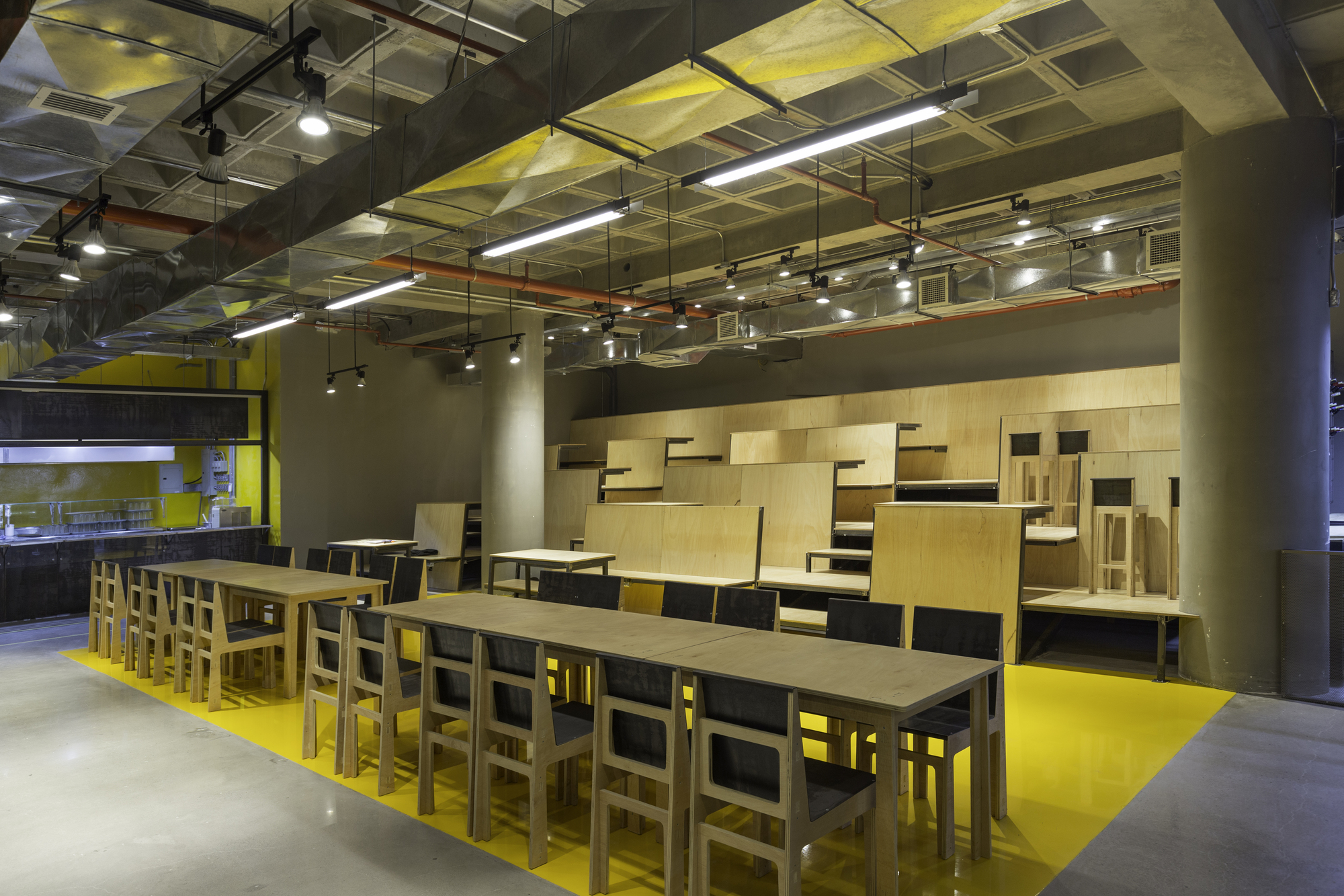 Long plywood table with black-seat chairs on a yellow floor strip, tiered plywood seating rising behind under suspended fluorescent lights