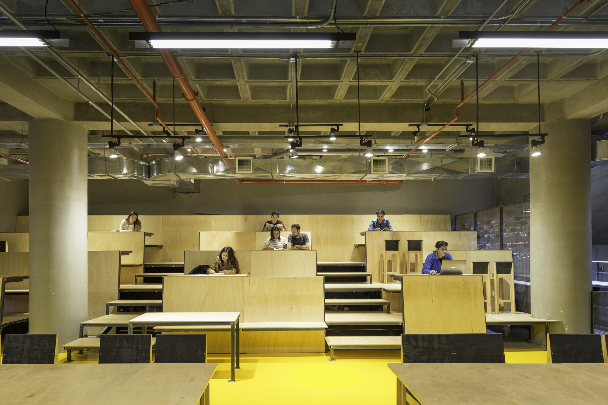 Tiered plywood seating platforms with students working on laptops between concrete columns beneath exposed orange pipes and a waffle slab