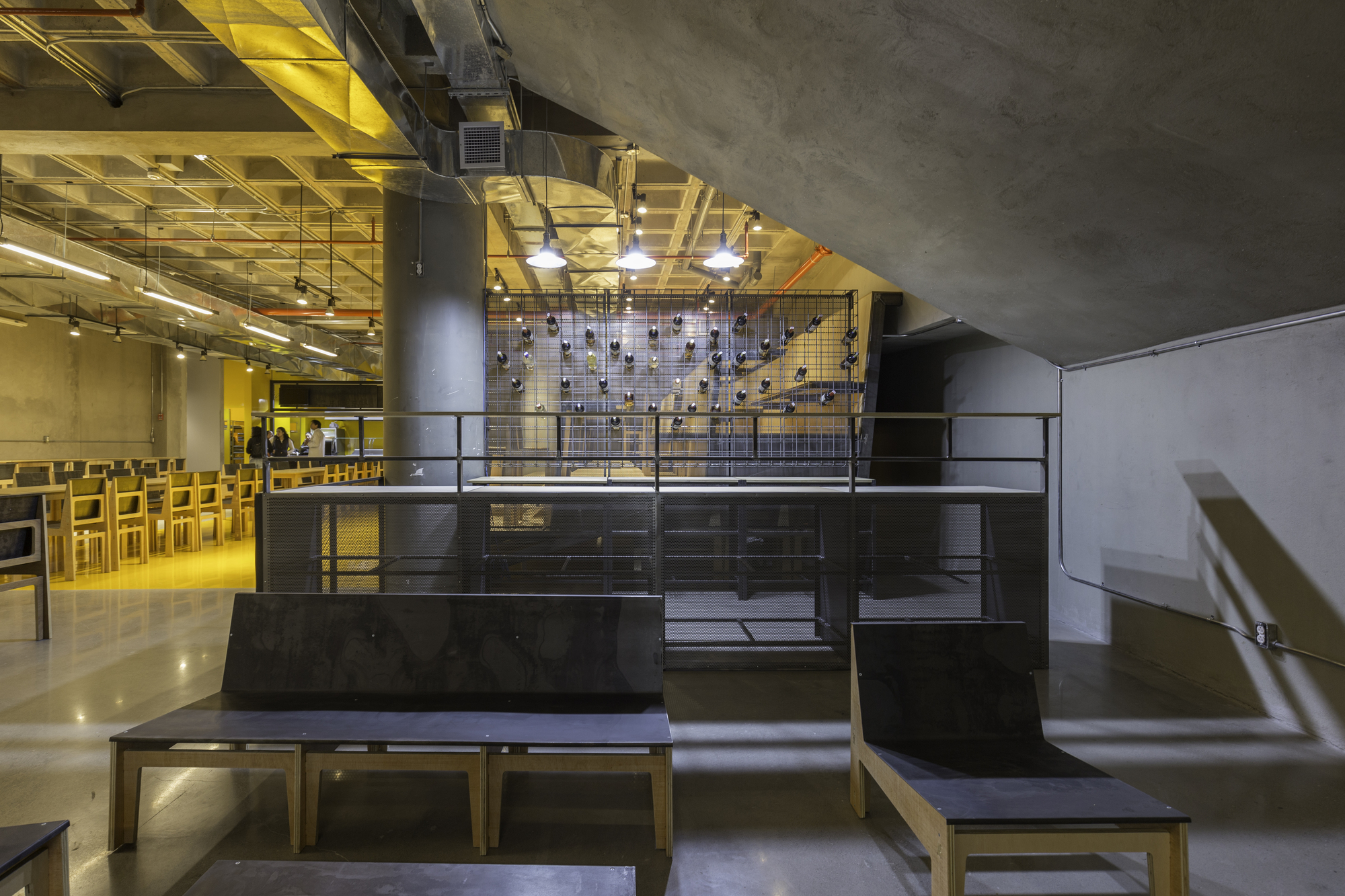 Black benches with timber legs in front of a mesh-backed platform holding hanging dark objects, beneath a sloping concrete underside
