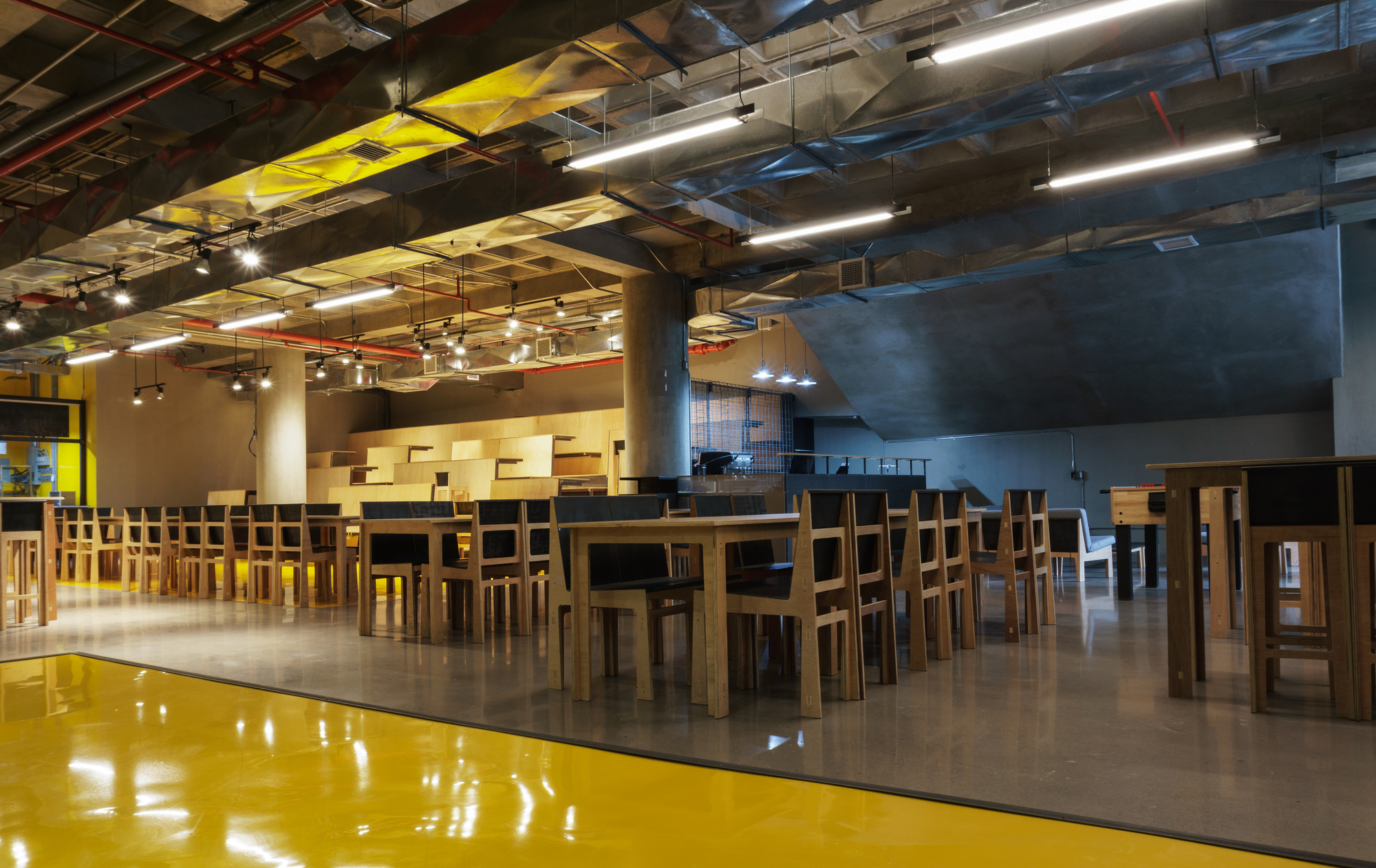 Wooden tables and chairs on a polished concrete floor beside a reflective yellow floor, with faceted metal ducts overhead