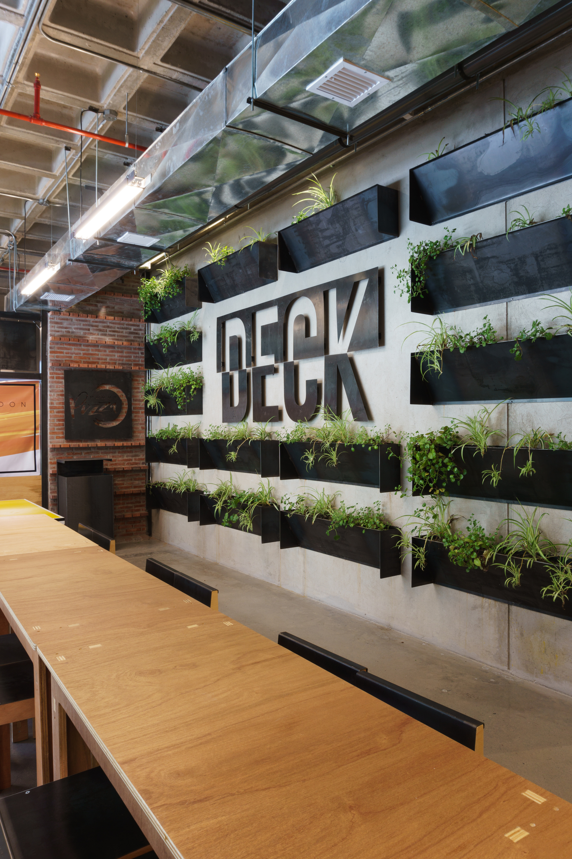 Concrete wall with oversized cut-out letters and rows of black steel planters, seen past a long plywood communal table