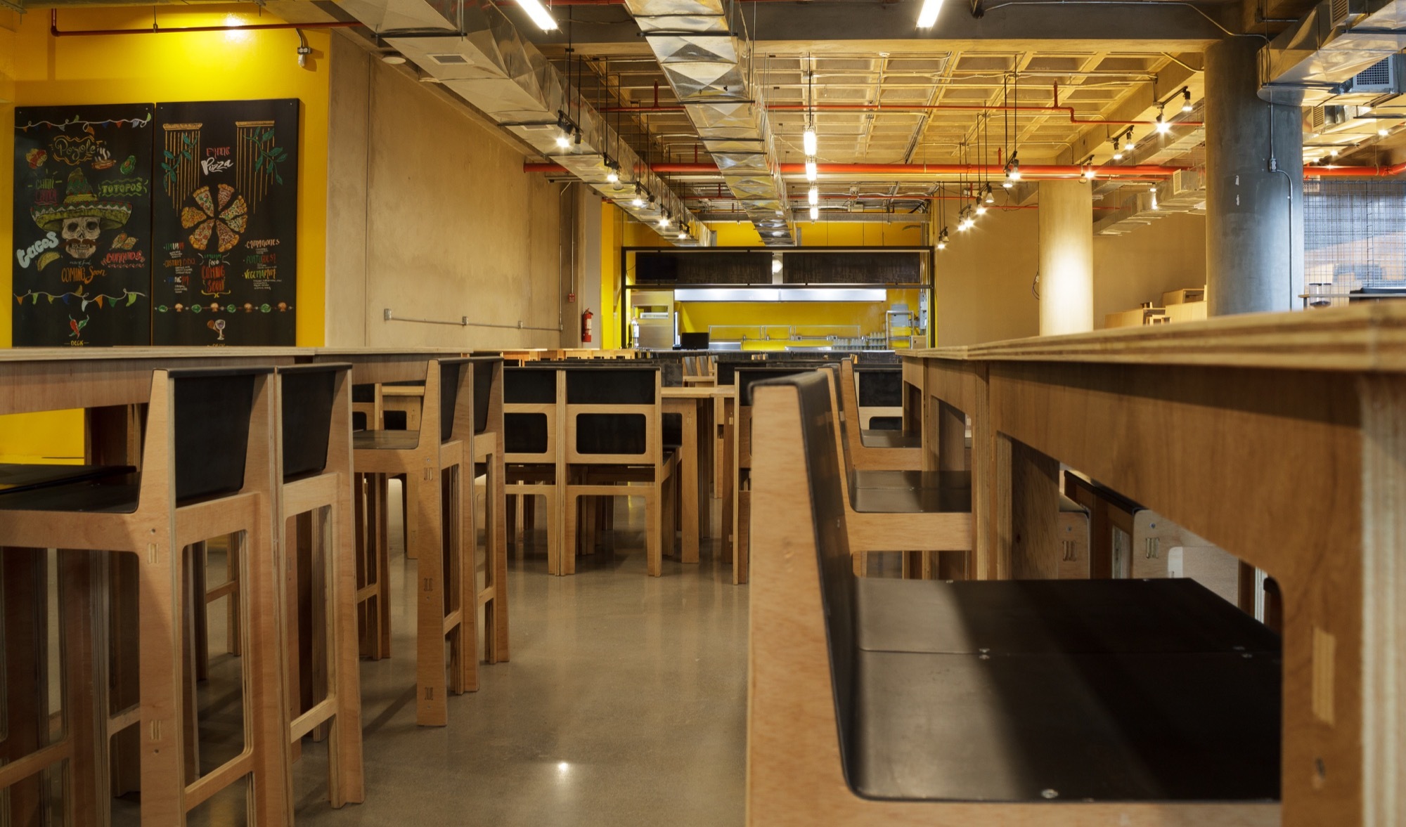 Row of tall plywood stools and tables leading toward a lit kitchen counter, with chalkboard art on the yellow wall
