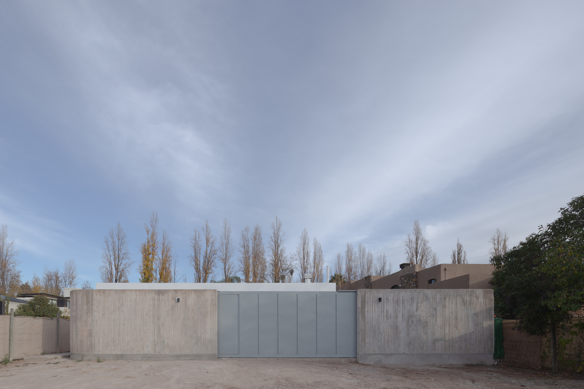 Street facade of board-marked concrete walls flanking a grey metal panel gate, with bare poplars behind under a streaked sky