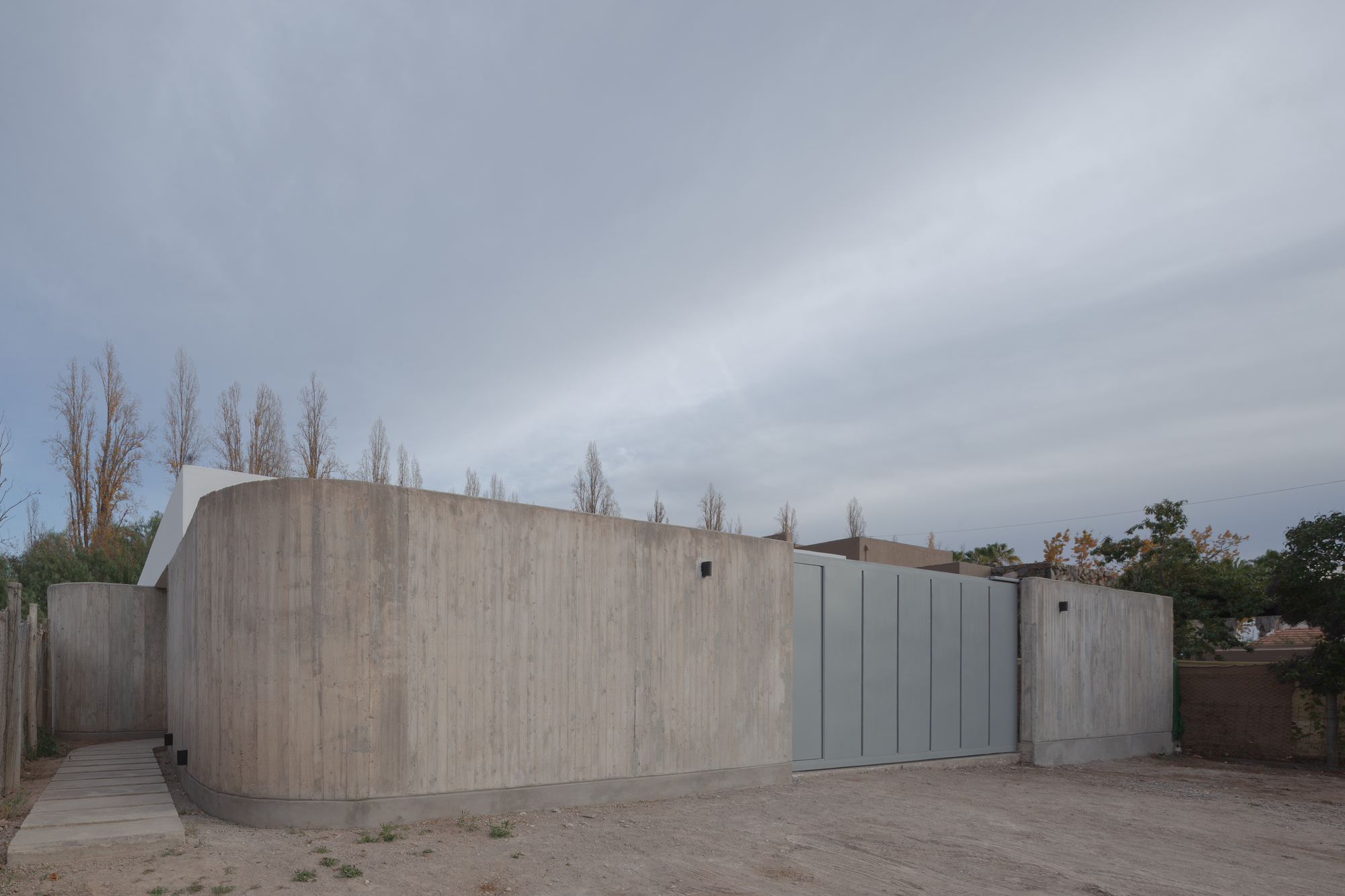 Curved board-formed concrete wall beside a grey panel gate and a paved stepping path under an overcast sky