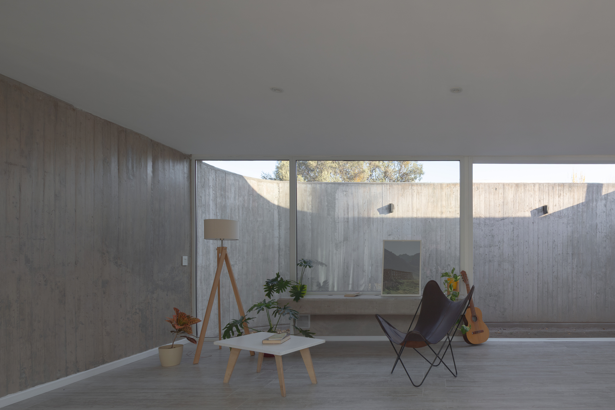 Living room with board-marked concrete wall, butterfly chair and tripod lamp facing a glazed wall onto a walled patio