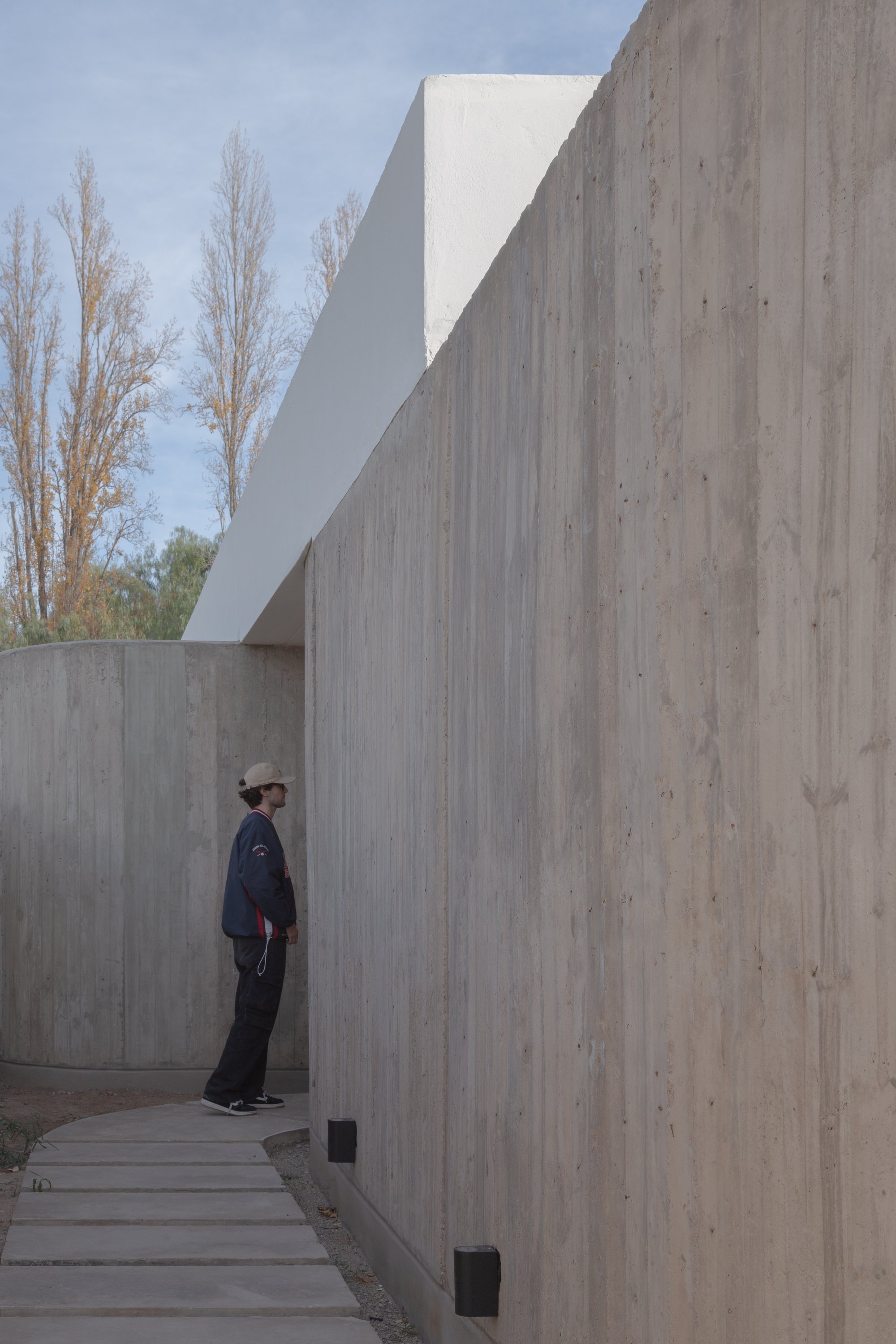 Person in a cap standing on a stepping-stone path beside a tall concrete wall, with yellow poplars behind