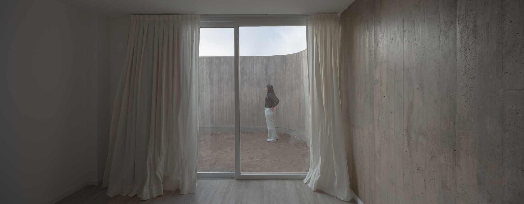 Bedroom with gathered white curtains framing a sliding glass door onto a curved concrete patio wall and a standing woman