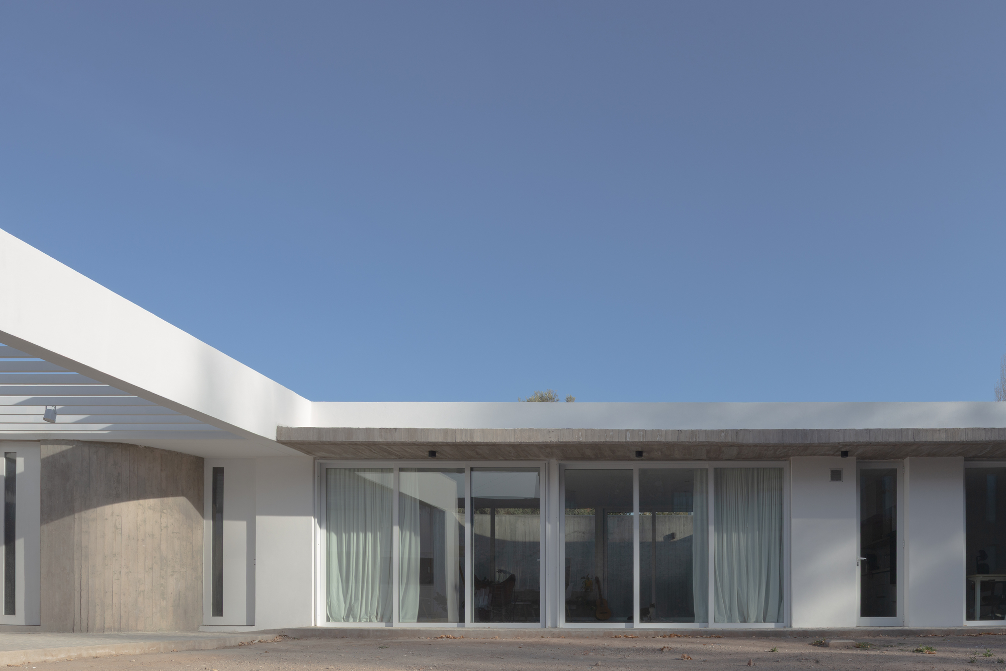 Low white-edged facade with floor-to-ceiling sliding glass, sheer curtains and a board-marked concrete soffit under a clear blue sky