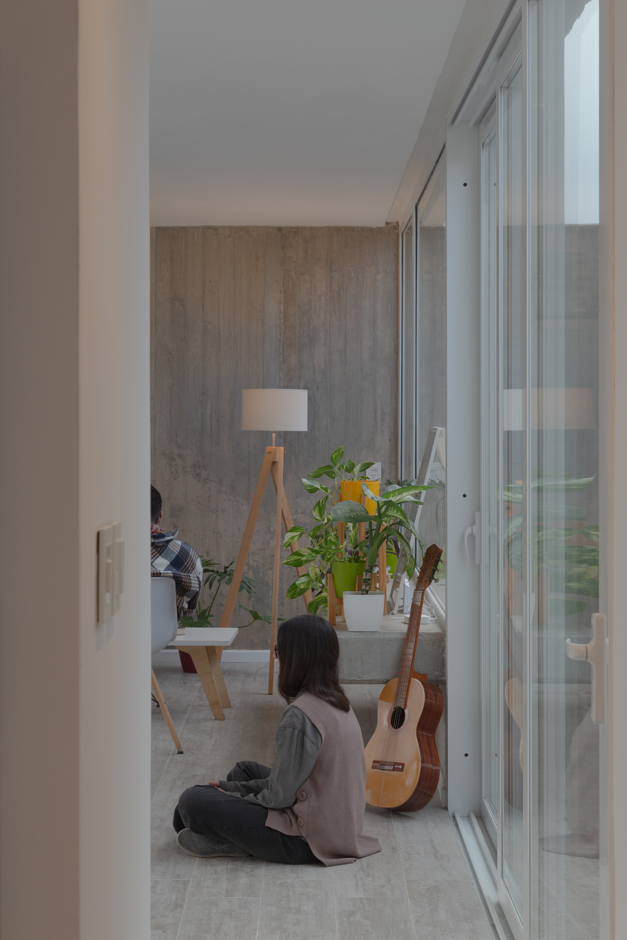 Living room corner with a seated woman, an acoustic guitar and potted plants beside a board-formed concrete wall