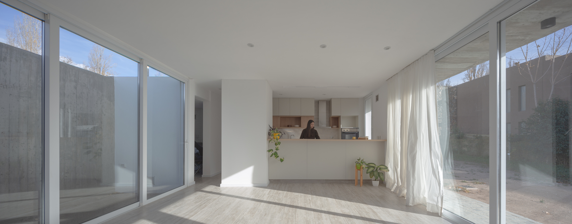 Open living and kitchen space with sliding glass walls, white curtains and a pale timber floor in afternoon sun