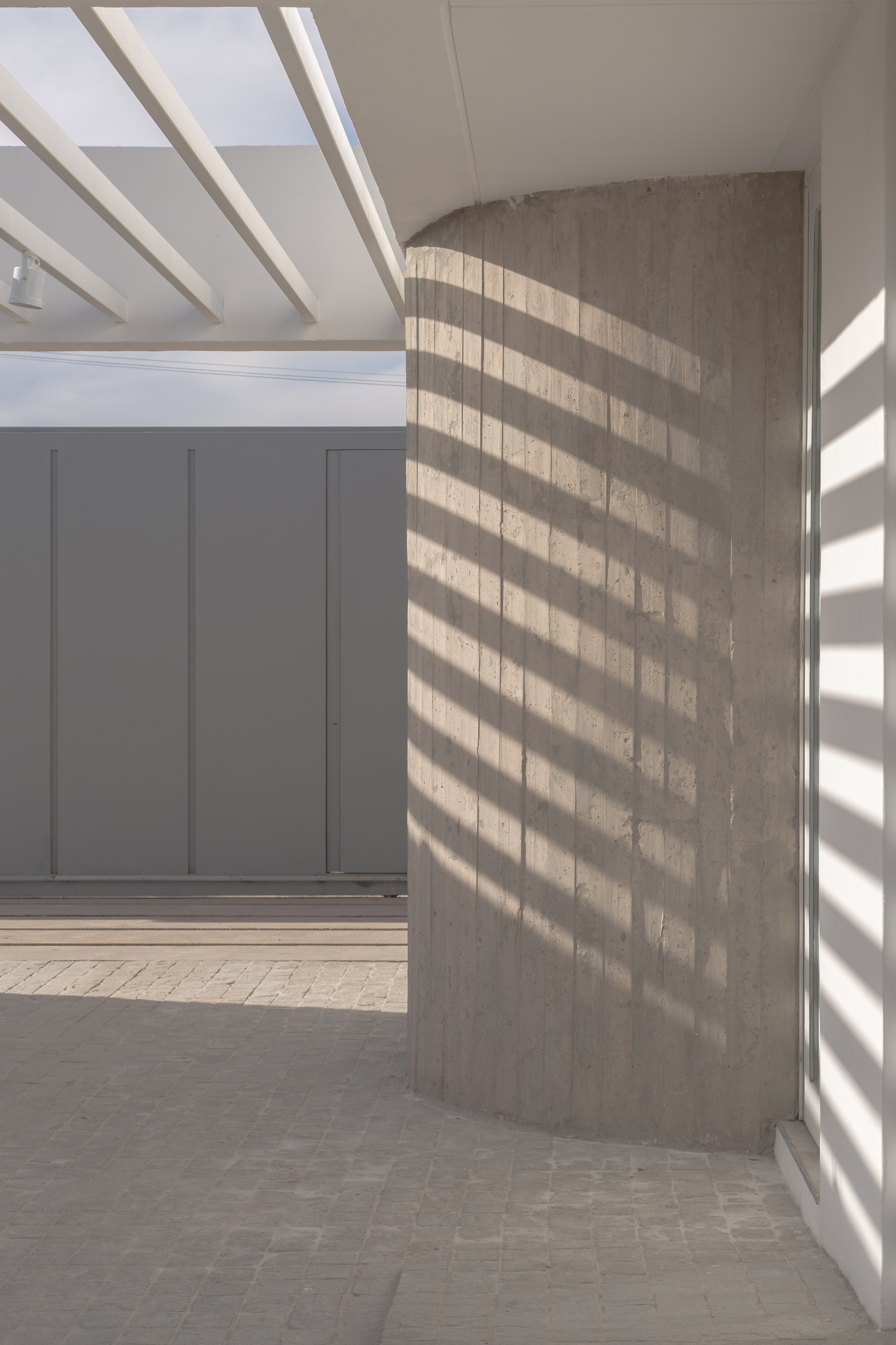 Curved board-formed concrete wall at a corner, striped by sunlight and shadow from the white pergola beams above