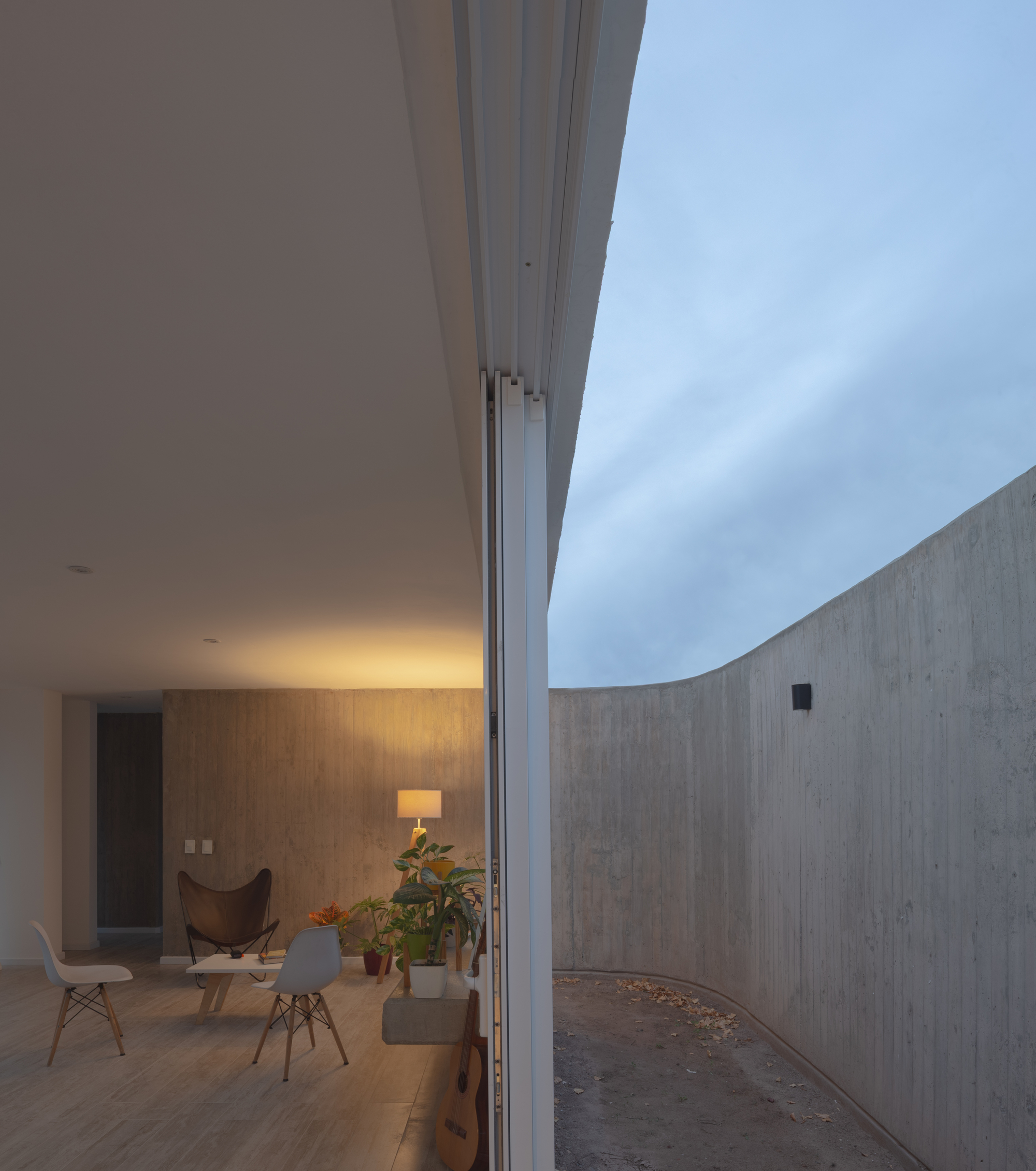 Dusk view from a glazed living room with warm lamplight toward a curving concrete courtyard wall and cloudy sky