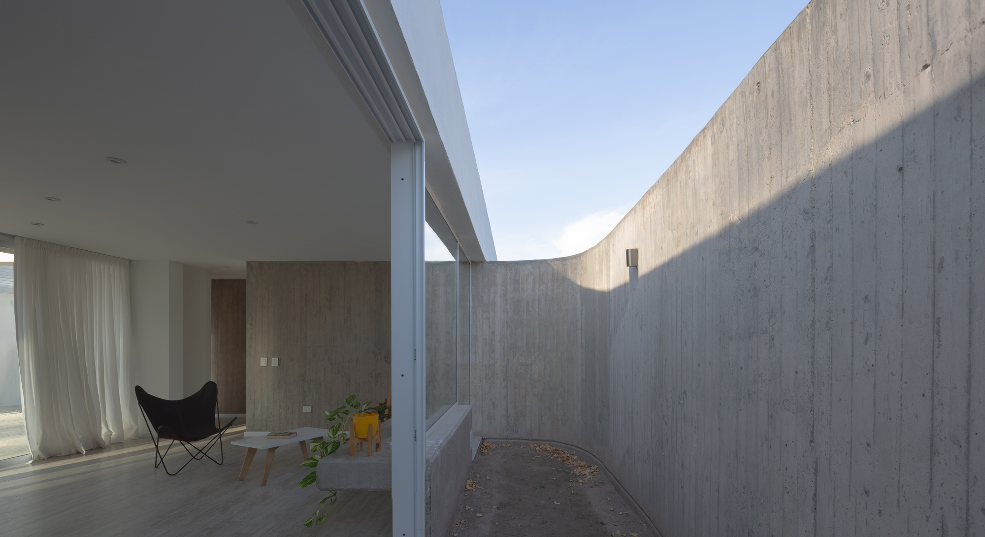 Curved concrete courtyard wall beside an open sliding glass door, with a butterfly chair inside under a bright sky