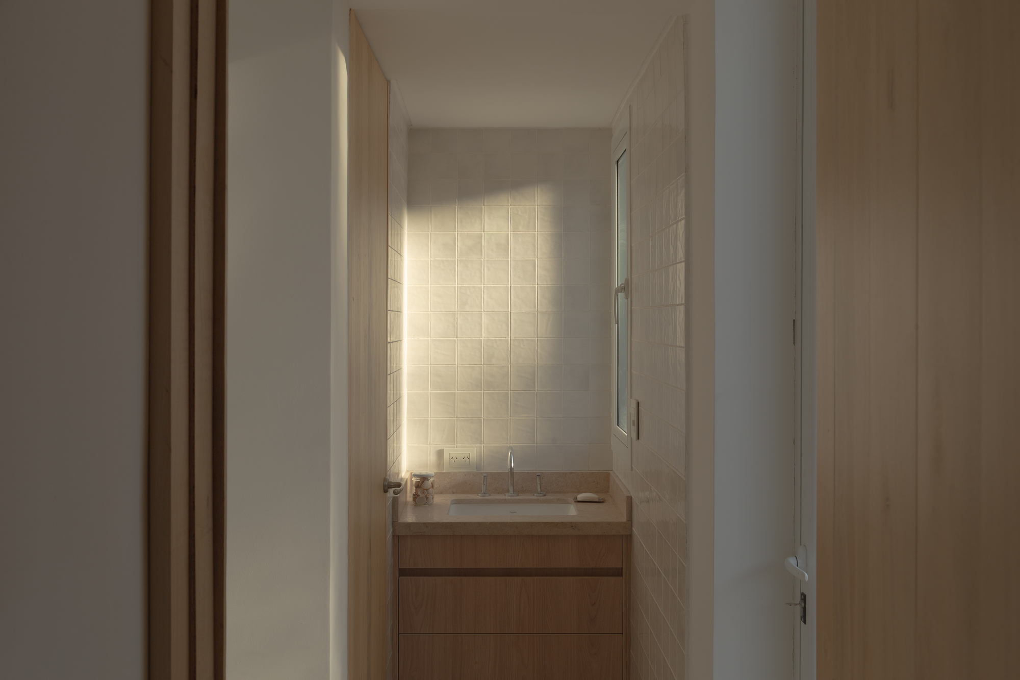 Small bathroom with a timber vanity and square handmade tile wall, with a patch of low sunlight falling across it