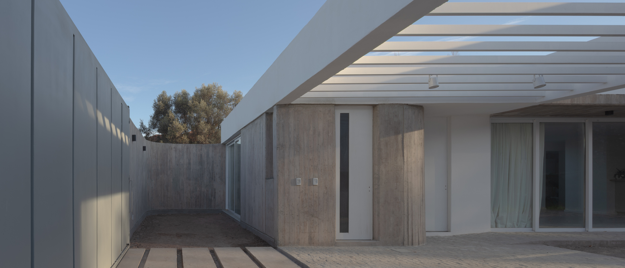 Entry court with a board-marked concrete curved volume and white pergola beams, under a clear blue afternoon sky