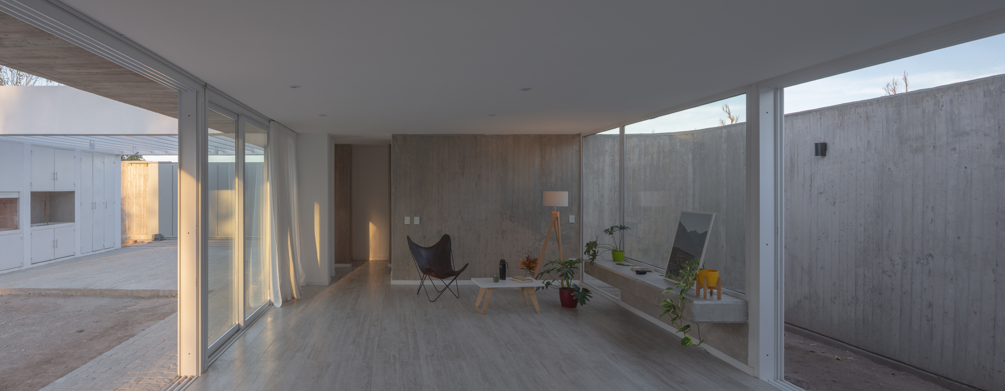 Open living space with a butterfly chair and low table between glass walls, framed by curved concrete courtyard walls at dusk