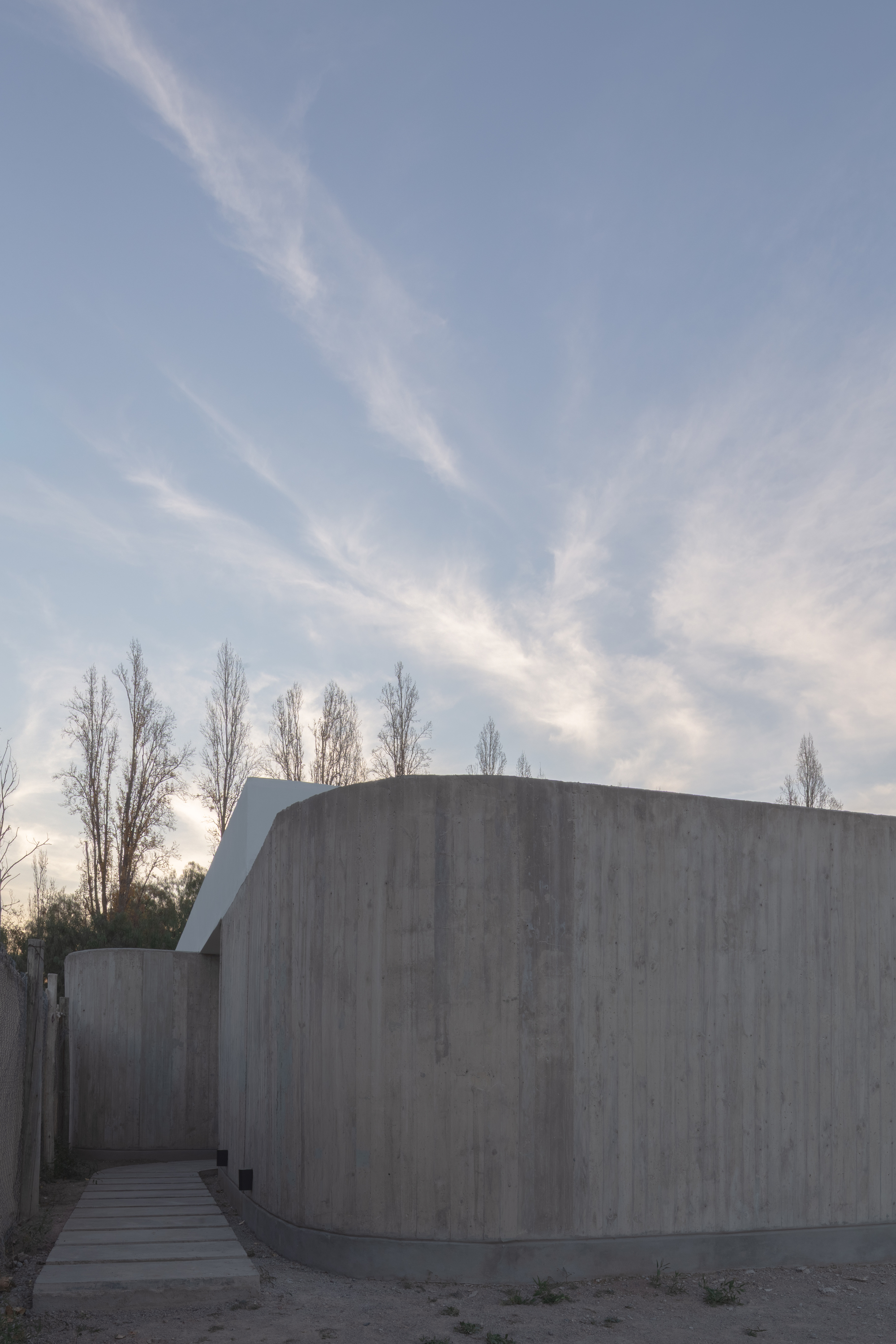 Curved board-formed concrete wall with a stepping-stone path, bare poplars behind and wispy clouds at dusk