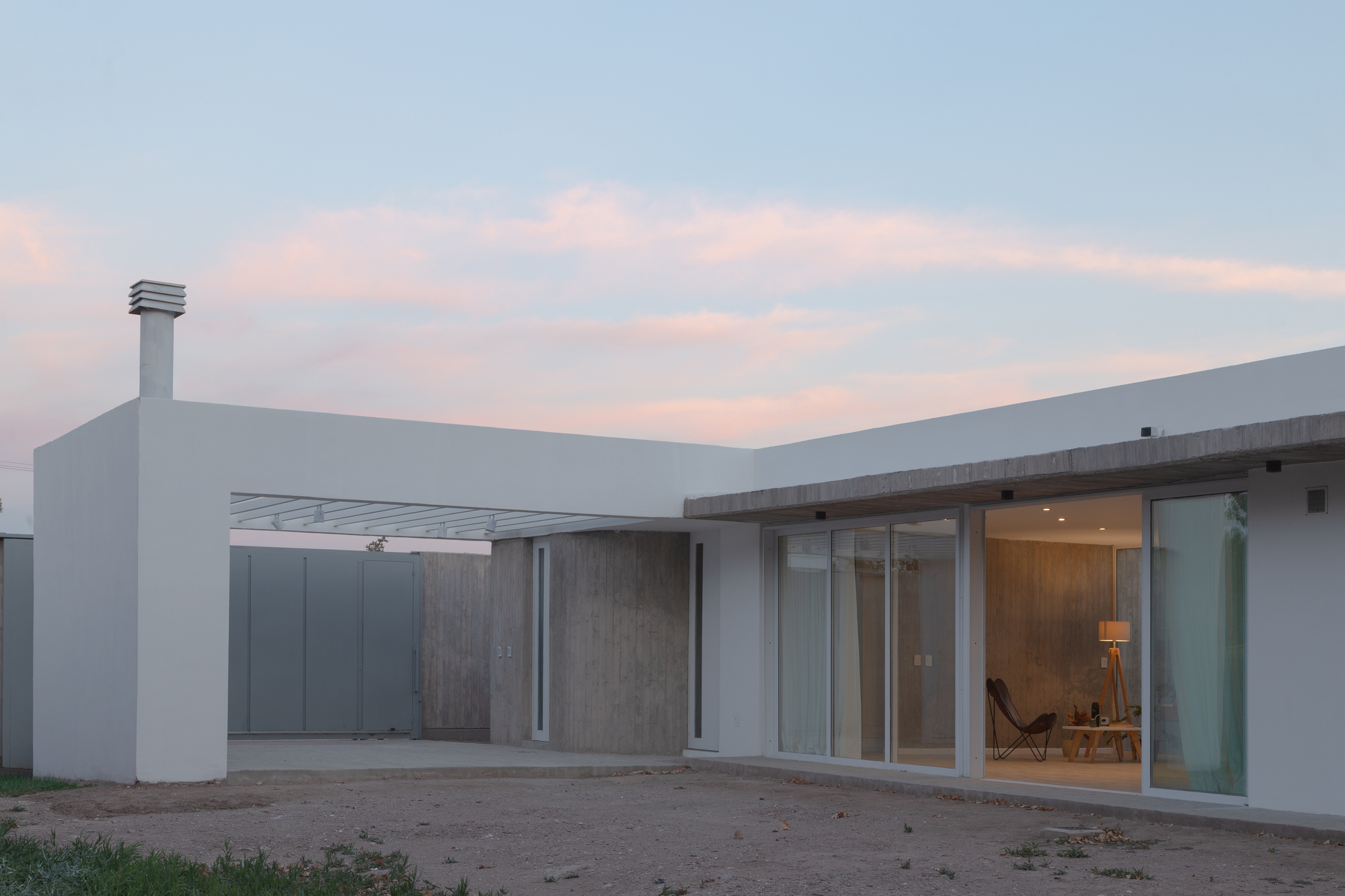 Single-storey white volume with a board-formed concrete wall and sliding glass doors, lit warmly inside under a pink dusk sky