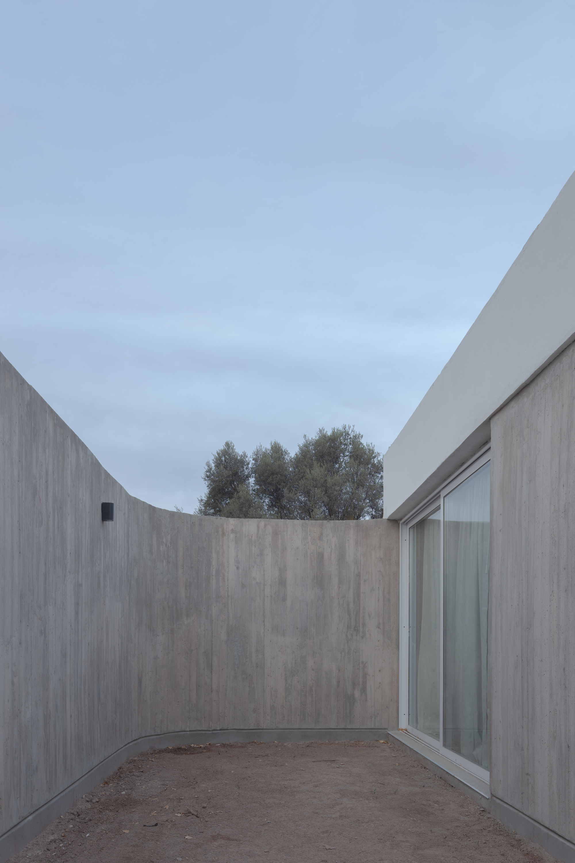 Narrow gravel court between a curved board-formed concrete wall and a glazed sliding door, with olive trees beyond under an overcast sky