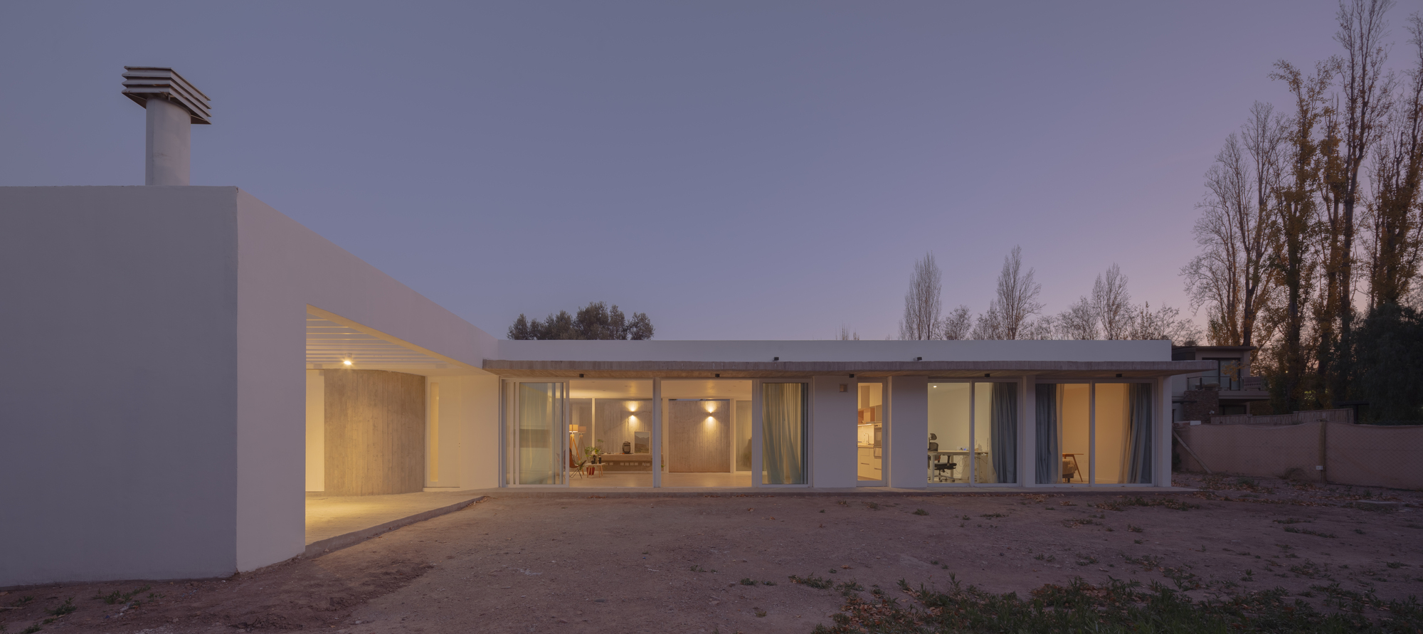 Wide view of the low white house with its chimney vent and glowing glazed living room at dusk, poplars standing bare to the right