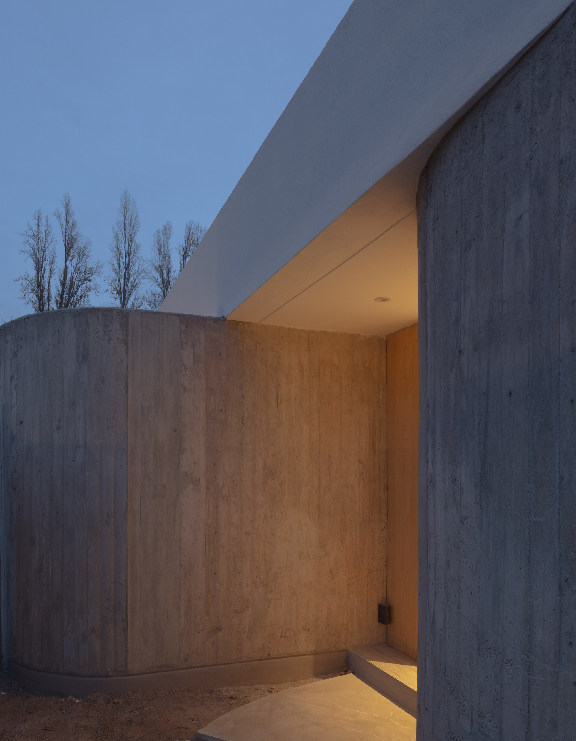 Entrance recess between two curved concrete walls, with a timber-lined reveal and a white roof edge overhead at dusk