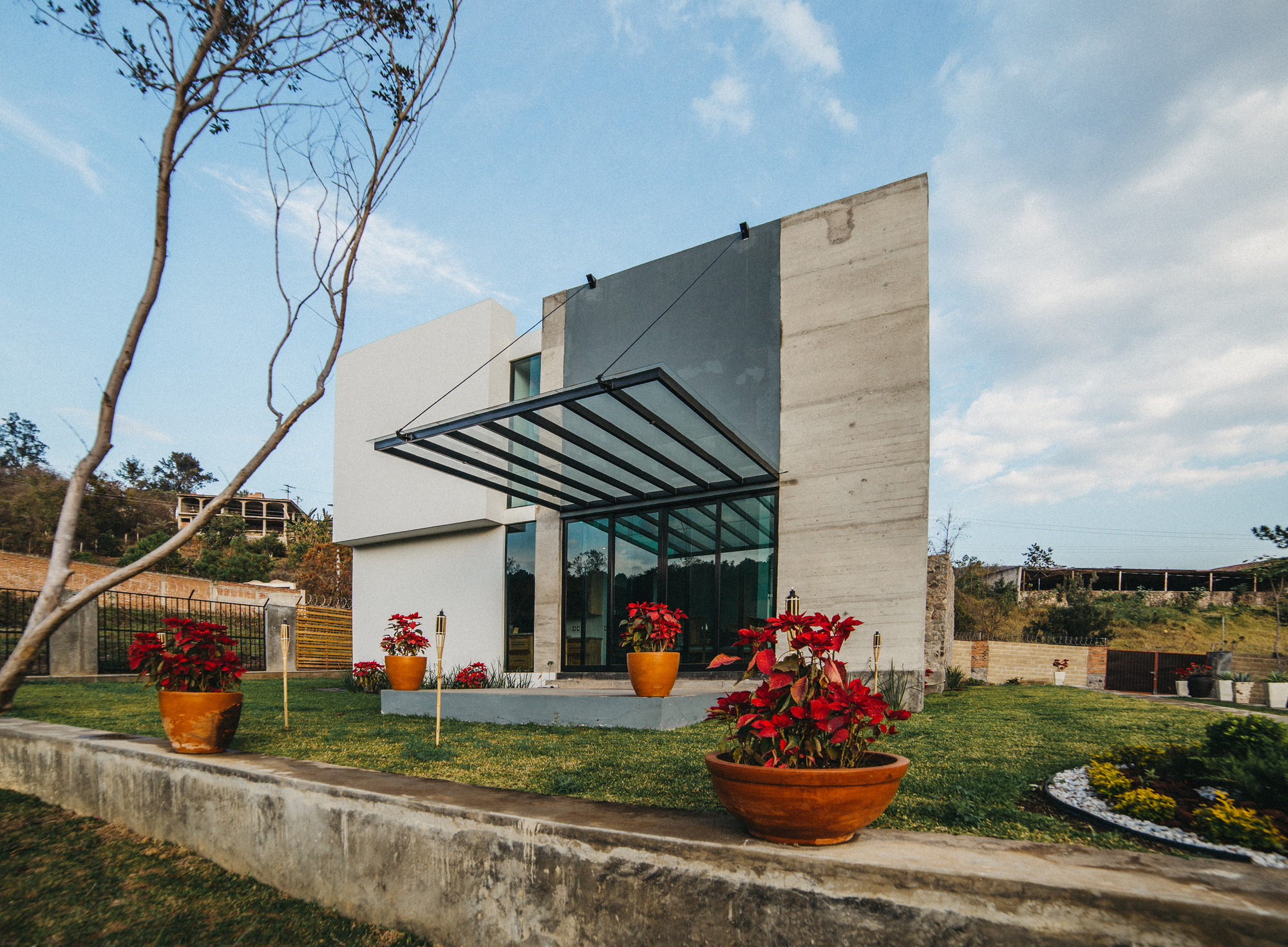 Glass canopy on steel rods over the entrance, with potted poinsettias on the lawn under a blue afternoon sky
