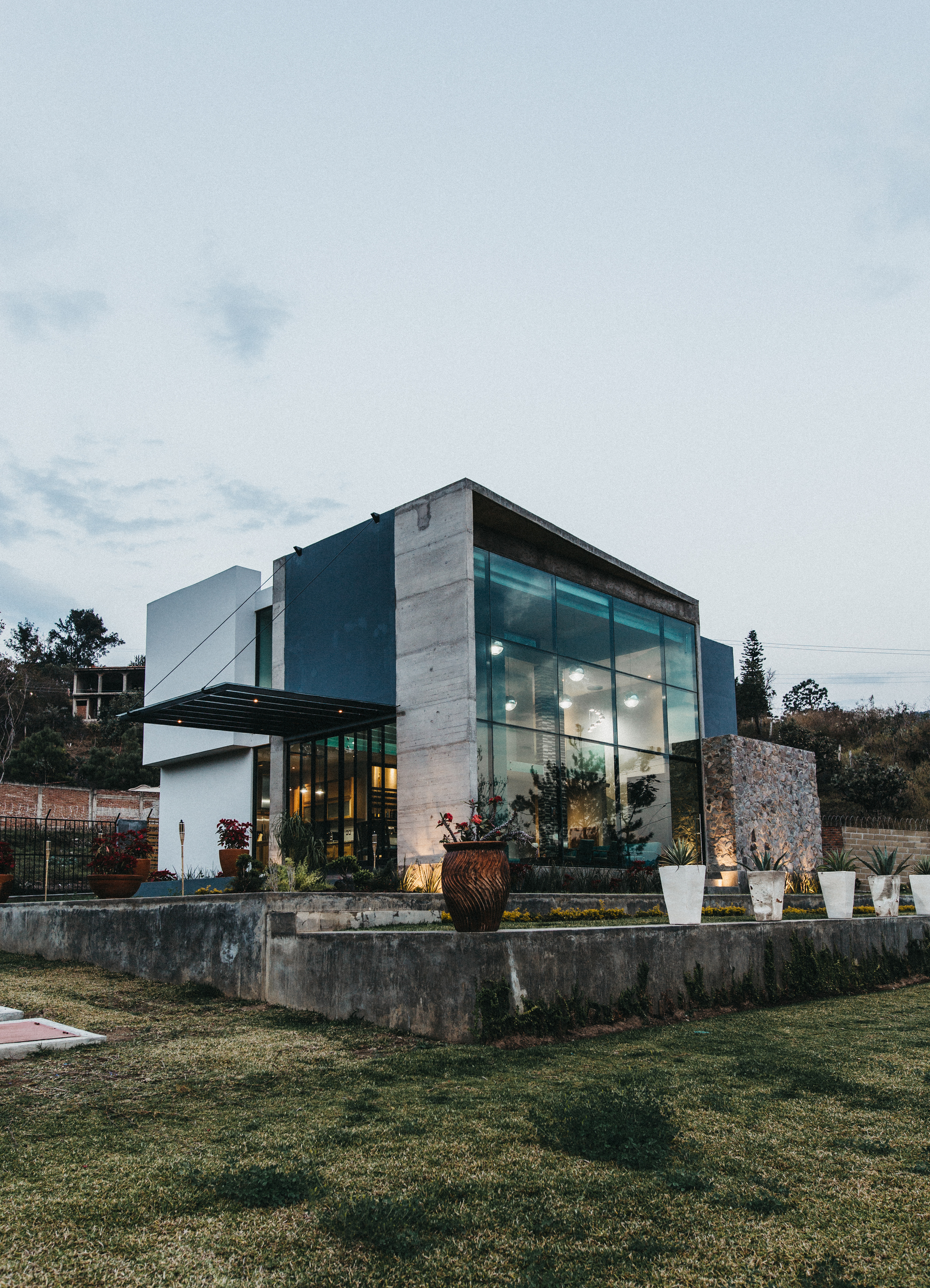 Dusk view of the lit glass volume, concrete pier and stone wall above a planted terrace with a clay urn