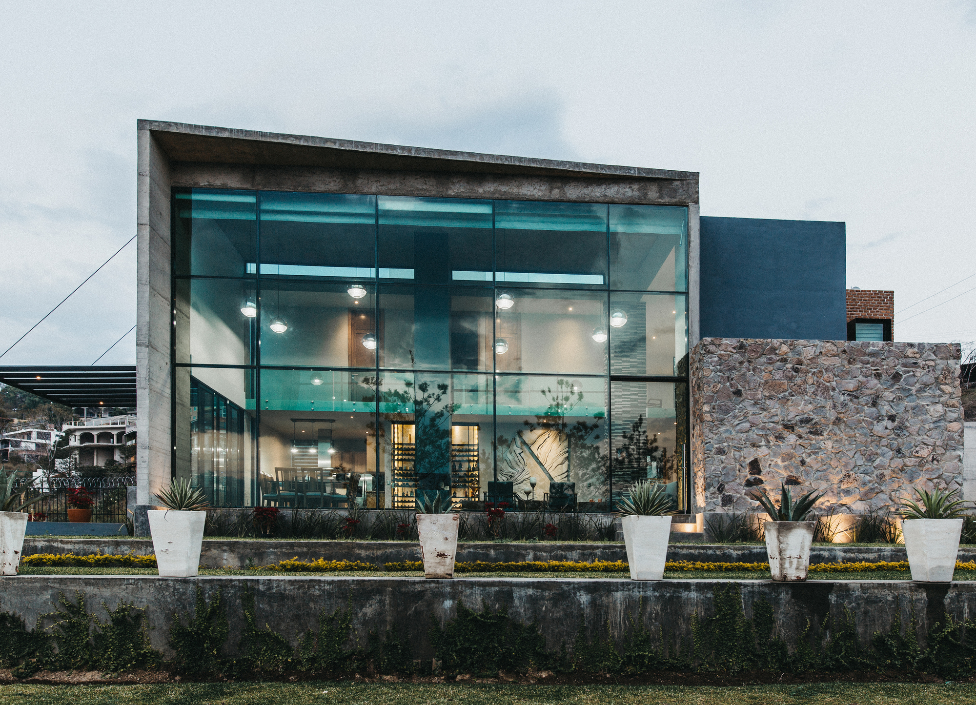 Front elevation of the full-height glass wall beside a rough stone wall, lit from within at twilight