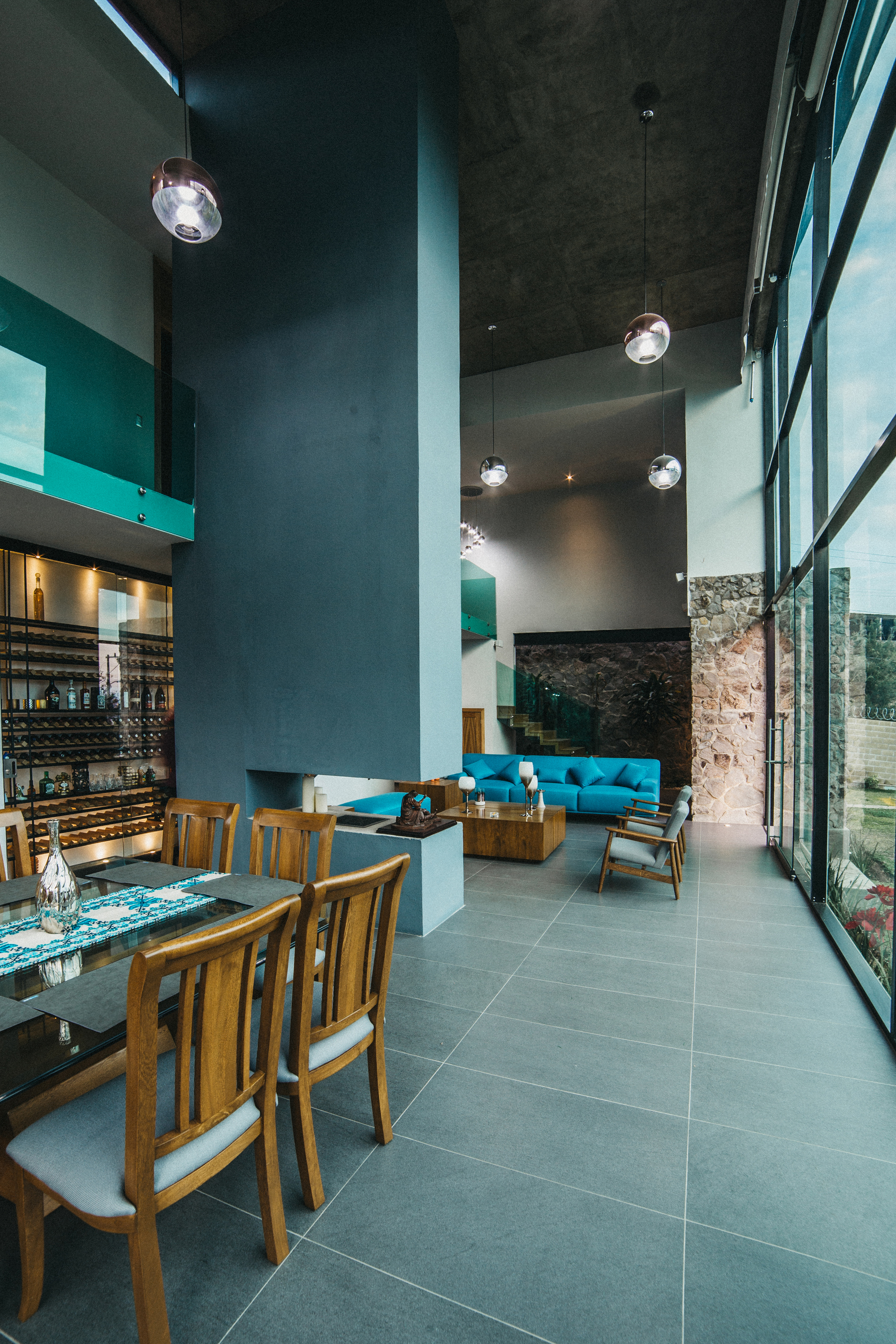 Double-height living room with teal blue sofa, wooden dining chairs, wine shelves and tall windows along grey tile floor