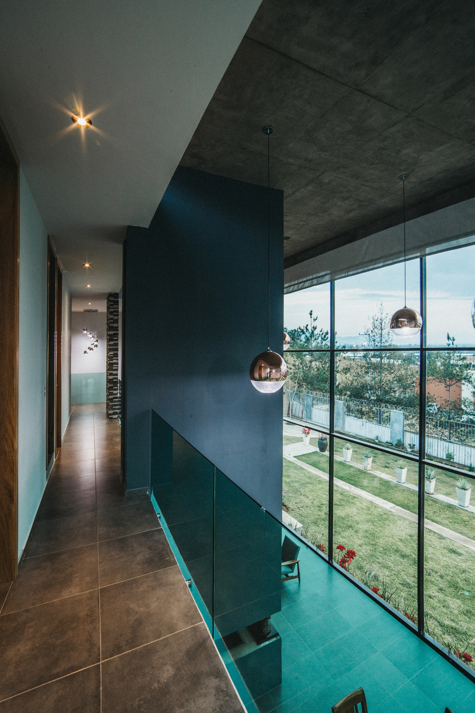 Upper hallway with a glass balustrade and copper pendant lamps beside a tall window overlooking a lawn in daylight