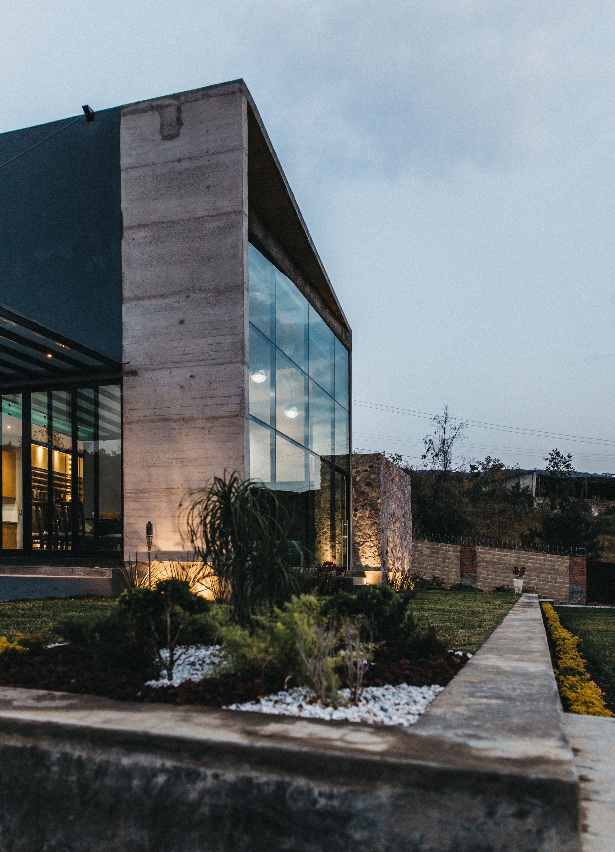 Board-formed concrete corner with a glass wall and stone pier, lit by garden lights at dusk