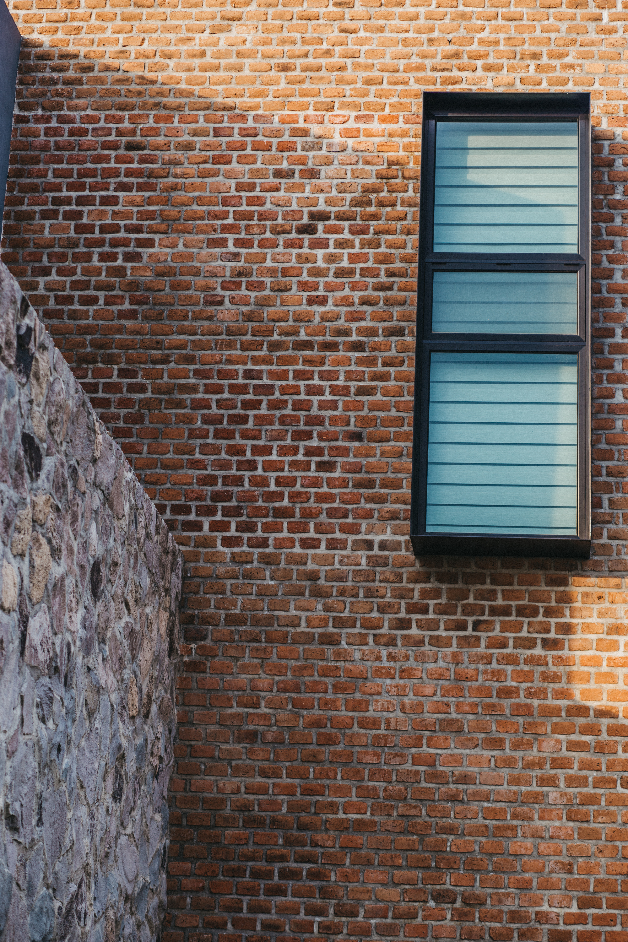 Red brick wall with a tall dark-framed window beside a rough stone wall, cut by afternoon shadow
