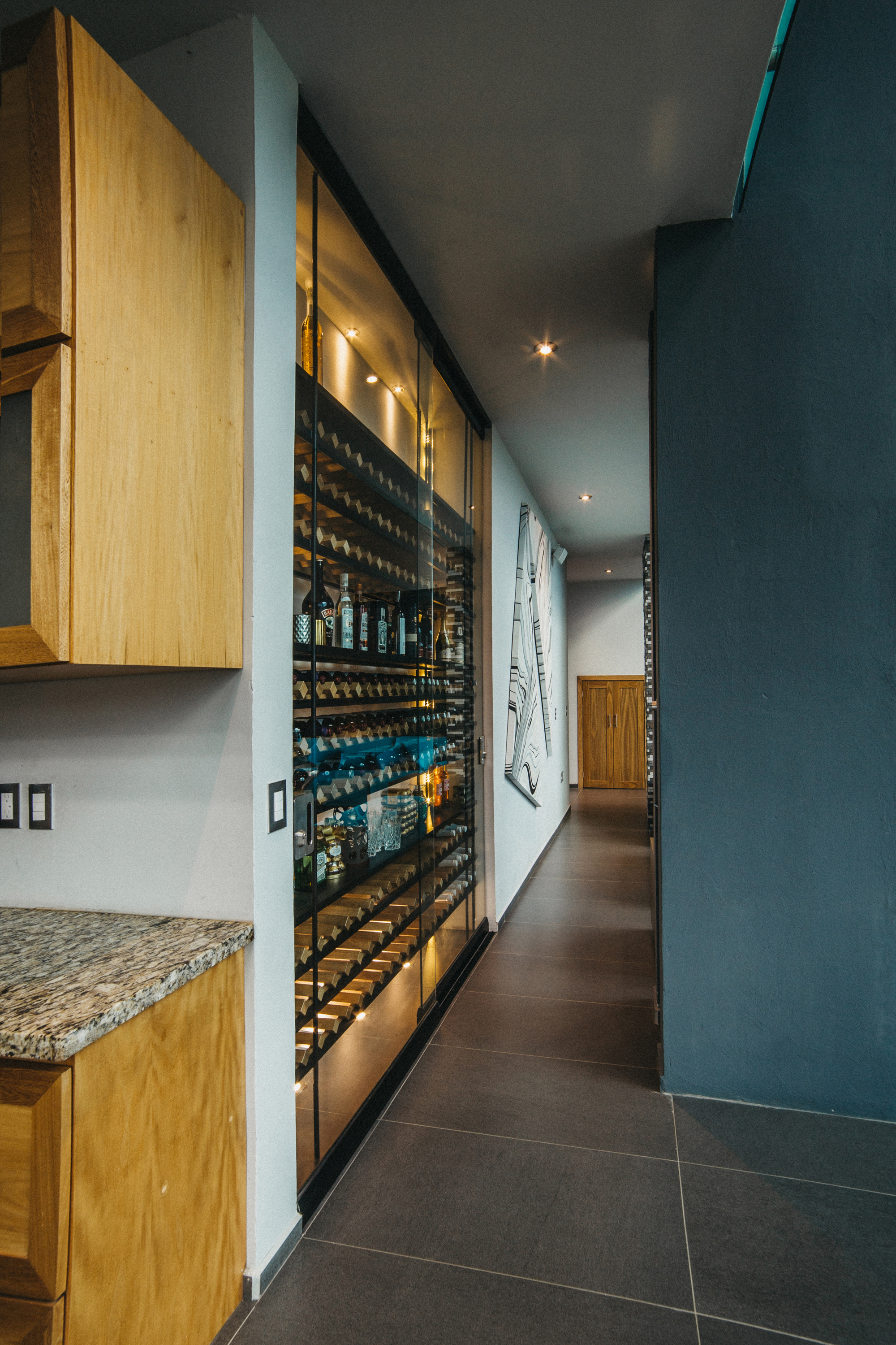 Glass-fronted wine cellar along a corridor with oak cabinets, granite counter and dark tile floor under recessed lights