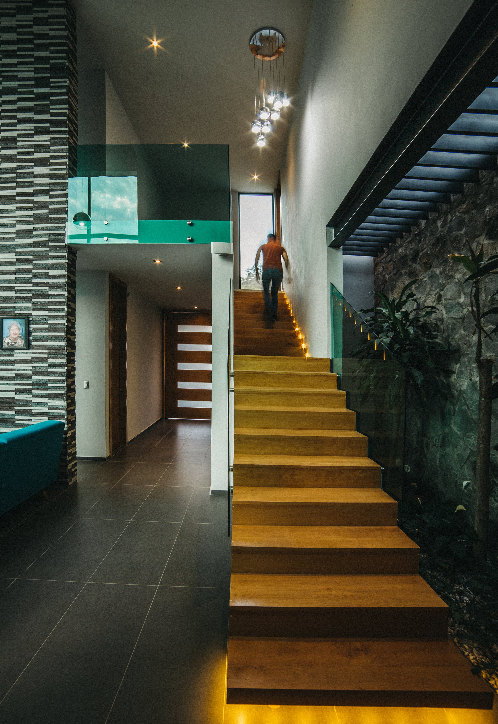 Timber staircase with lit risers rising past a stone wall and indoor planting as a person climbs