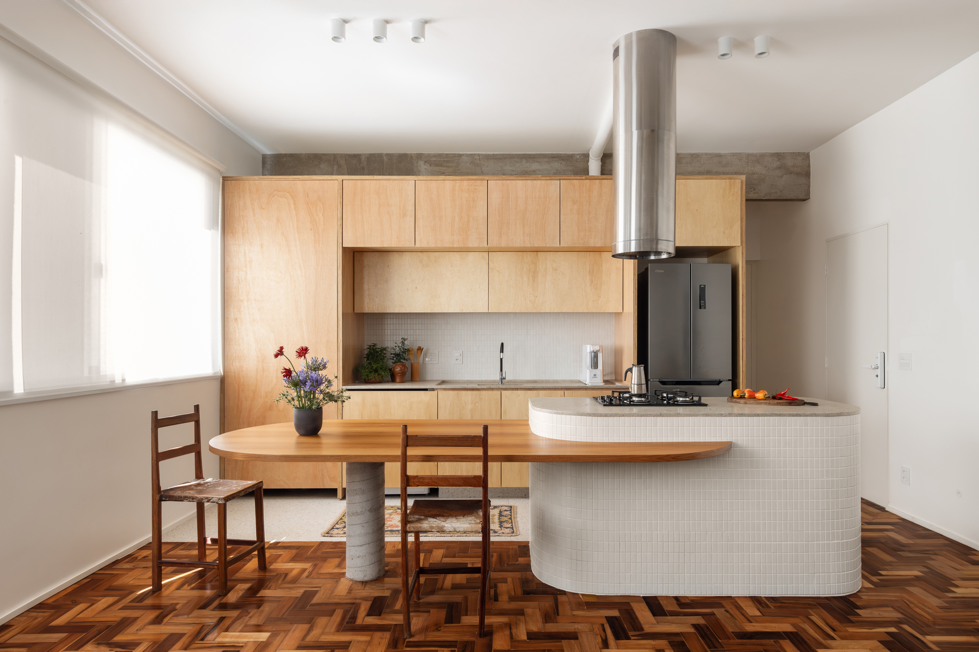 Kitchen island clad in white mosaic tile with a curved timber table extension, set on herringbone parquet in soft window light