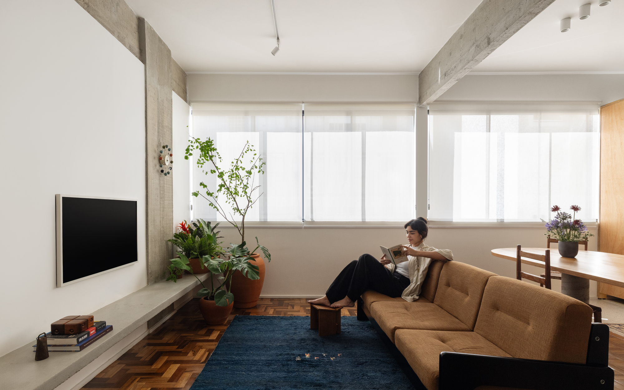 Woman reading on a tan sofa beside a concrete beam and potted plants, with daylight filtering through sheer roller blinds
