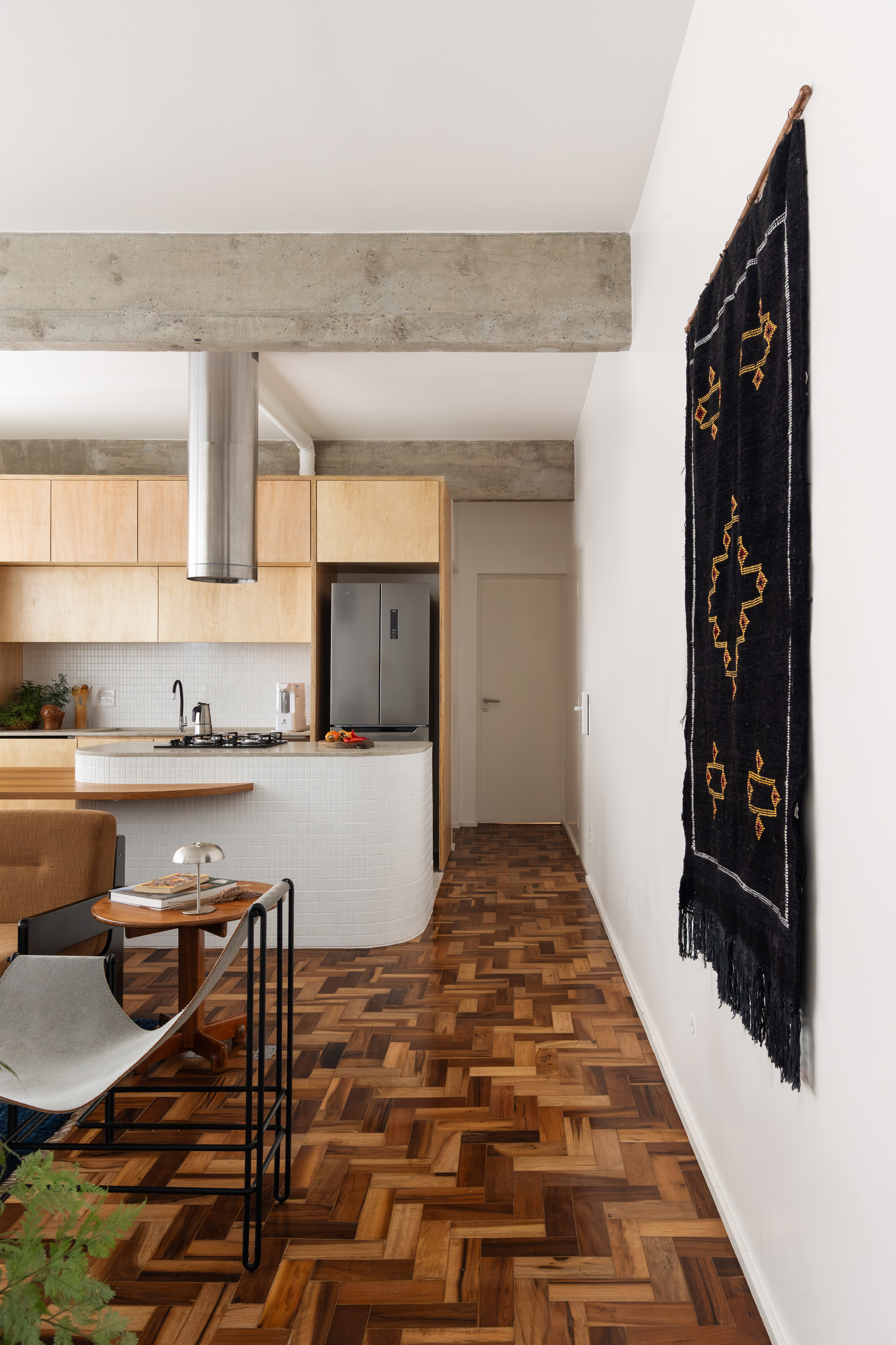 Hallway view along a black woven wall hanging toward the tiled island, with exposed concrete beams over herringbone parquet