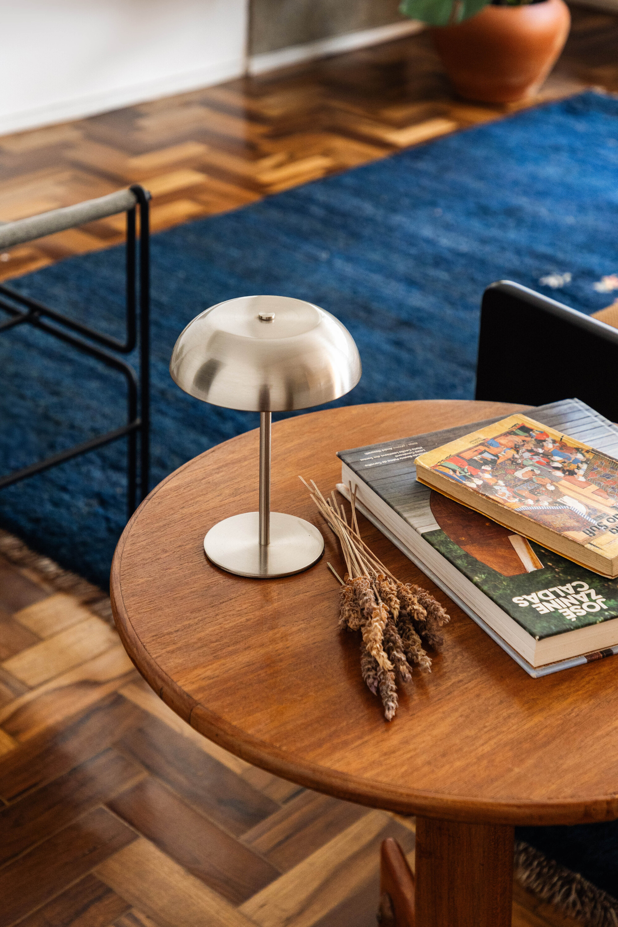 Round teak side table holding a brushed metal dome lamp, dried lavender stems and stacked books on a blue wool rug