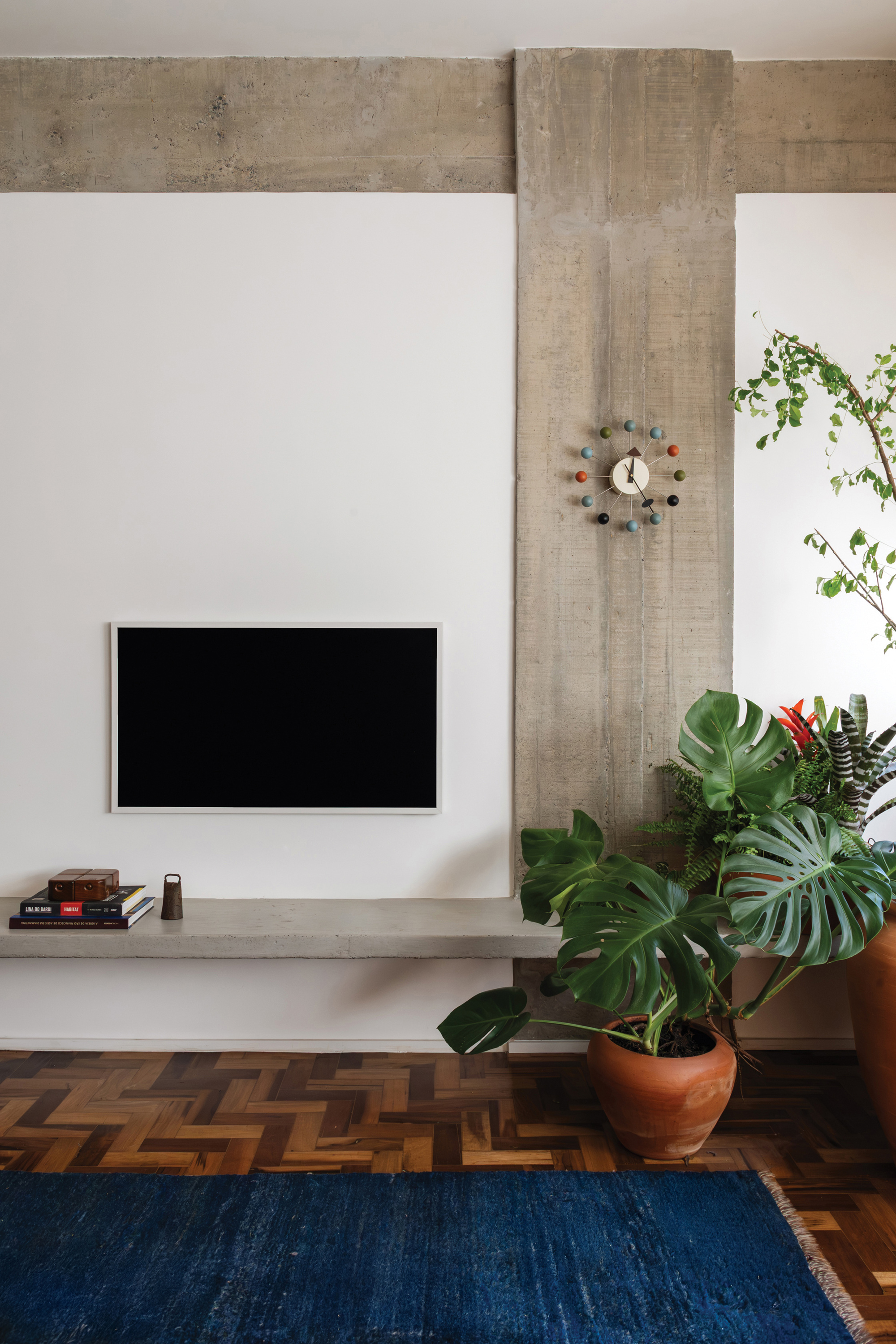Board-formed concrete column with a ball clock above a floating concrete shelf, a monstera in a terracotta pot beside it