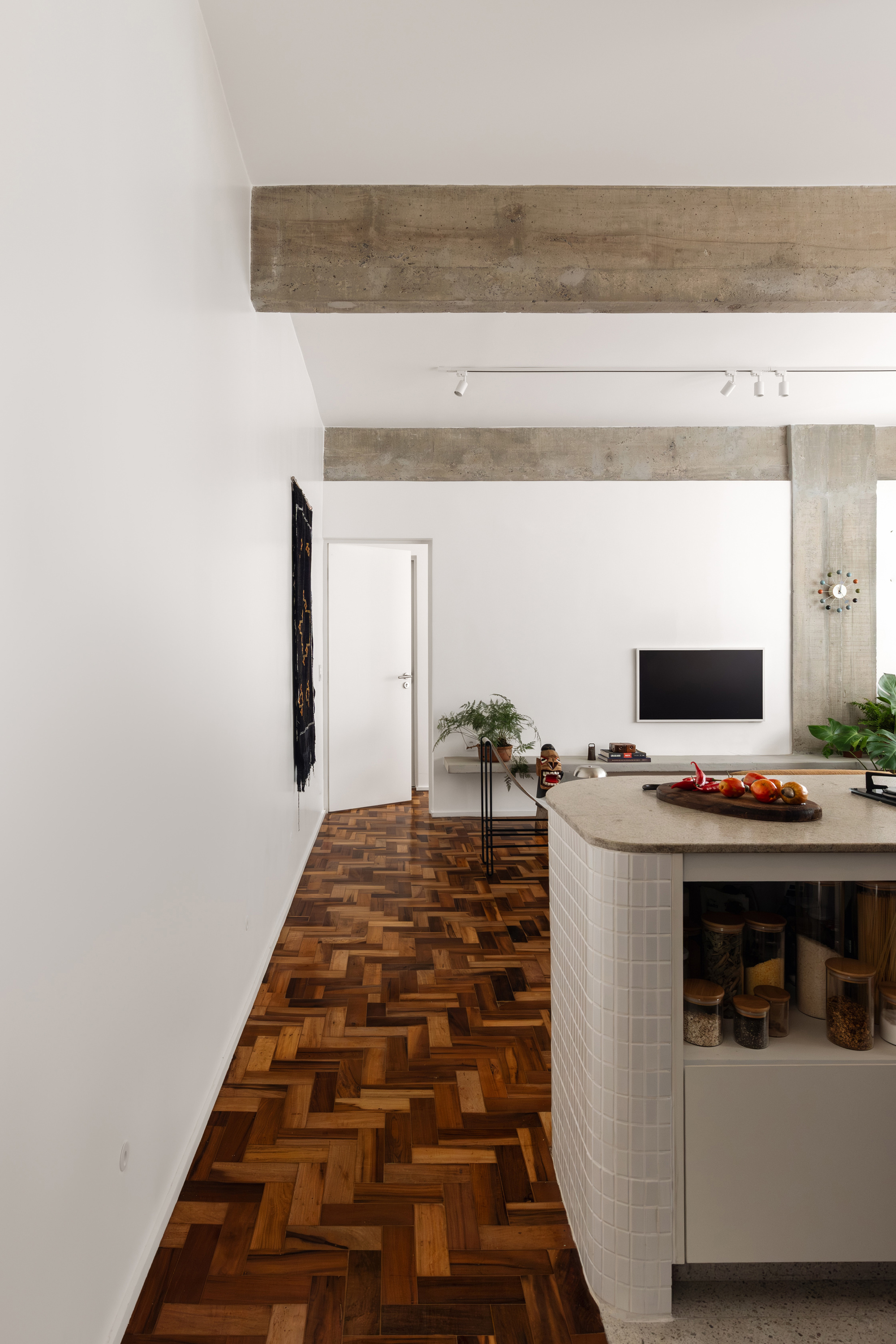 Herringbone wood floor running past a tiled kitchen island toward a concrete beam and wall-mounted television