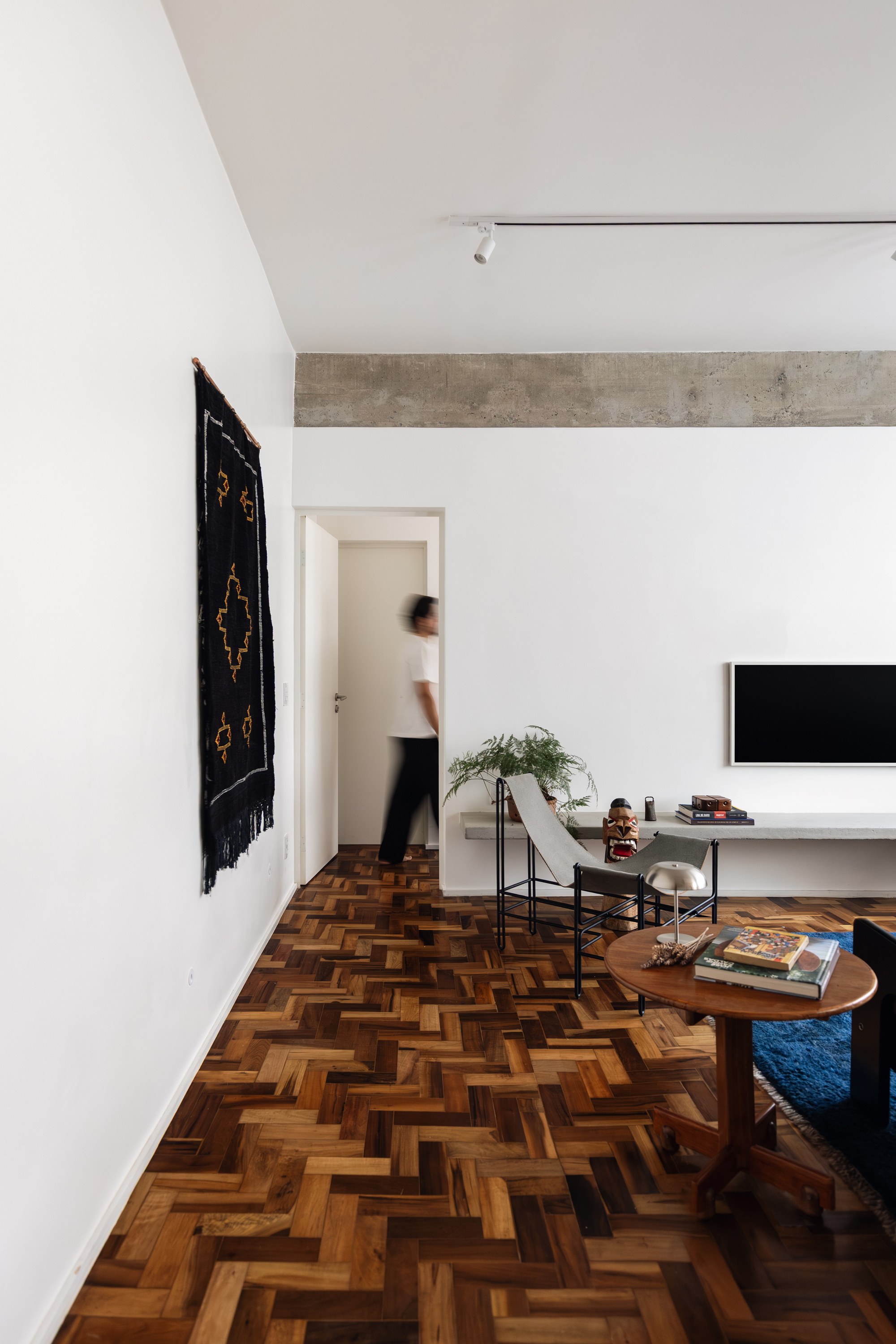 Person walking through a doorway beside a black woven wall hanging, with herringbone parquet and a round side table in the foreground