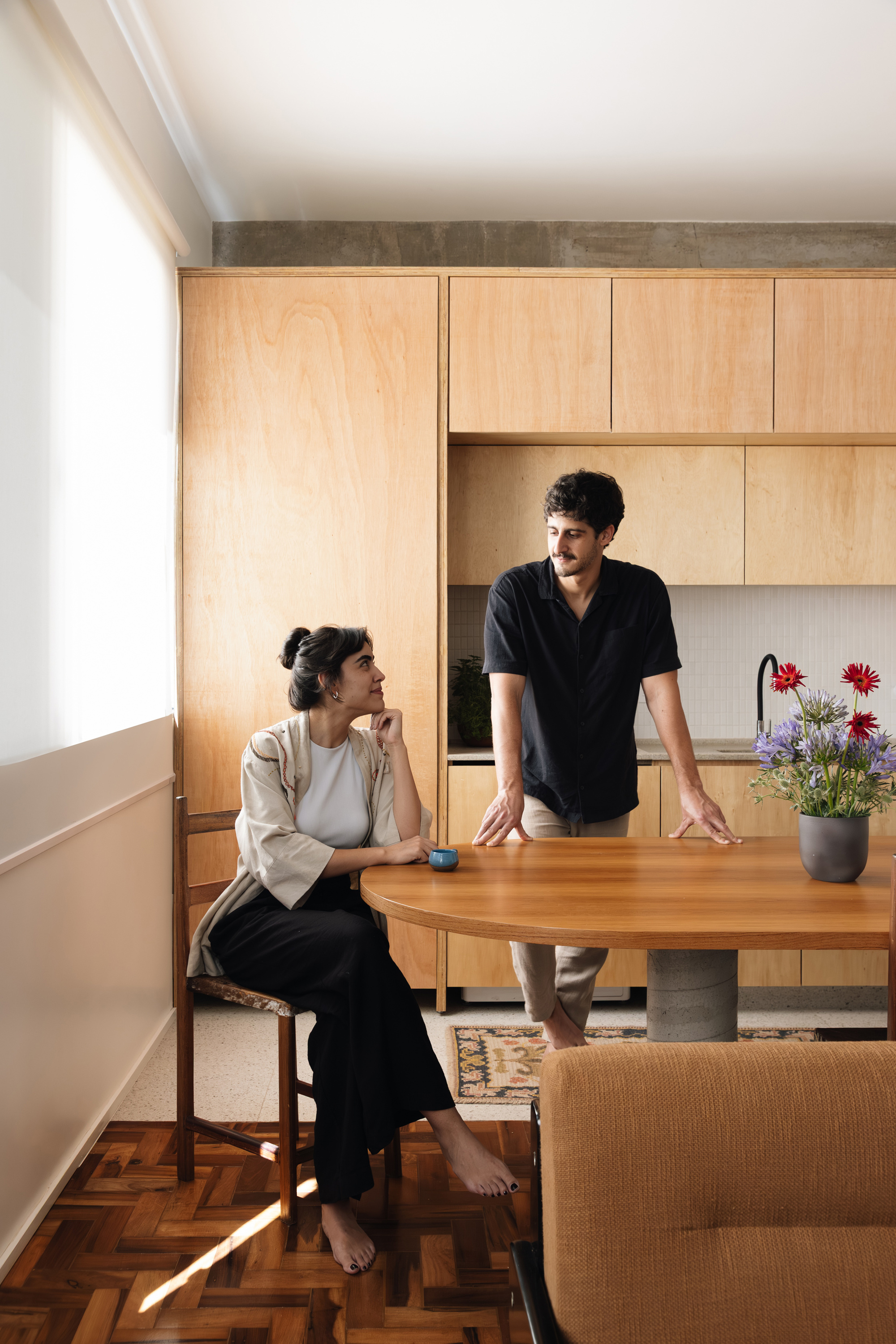 Seated woman and standing man at an oval timber table before plywood kitchen cabinets, with soft daylight from a side window