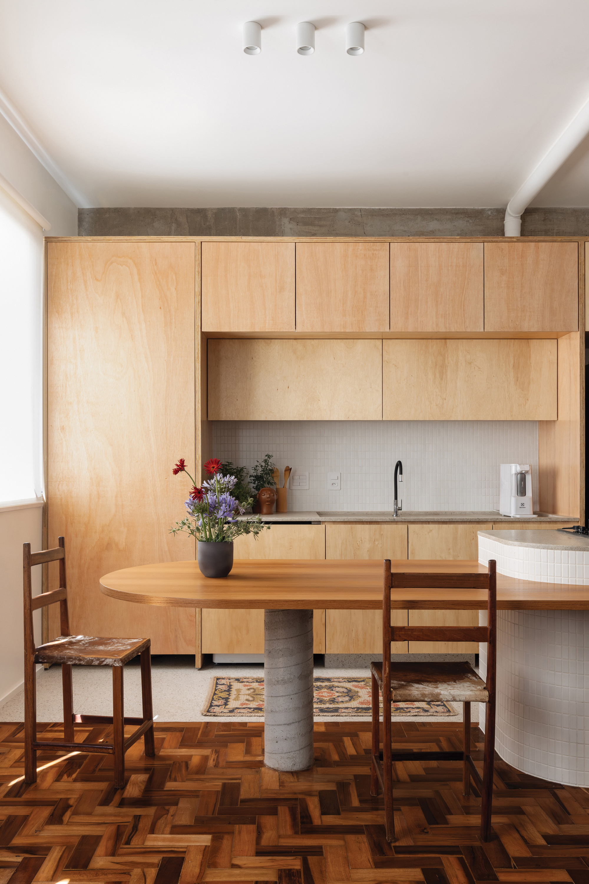 Plywood kitchen wall with an oval oak table on a concrete pedestal, parquet floor and a ladder-back chair in daylight