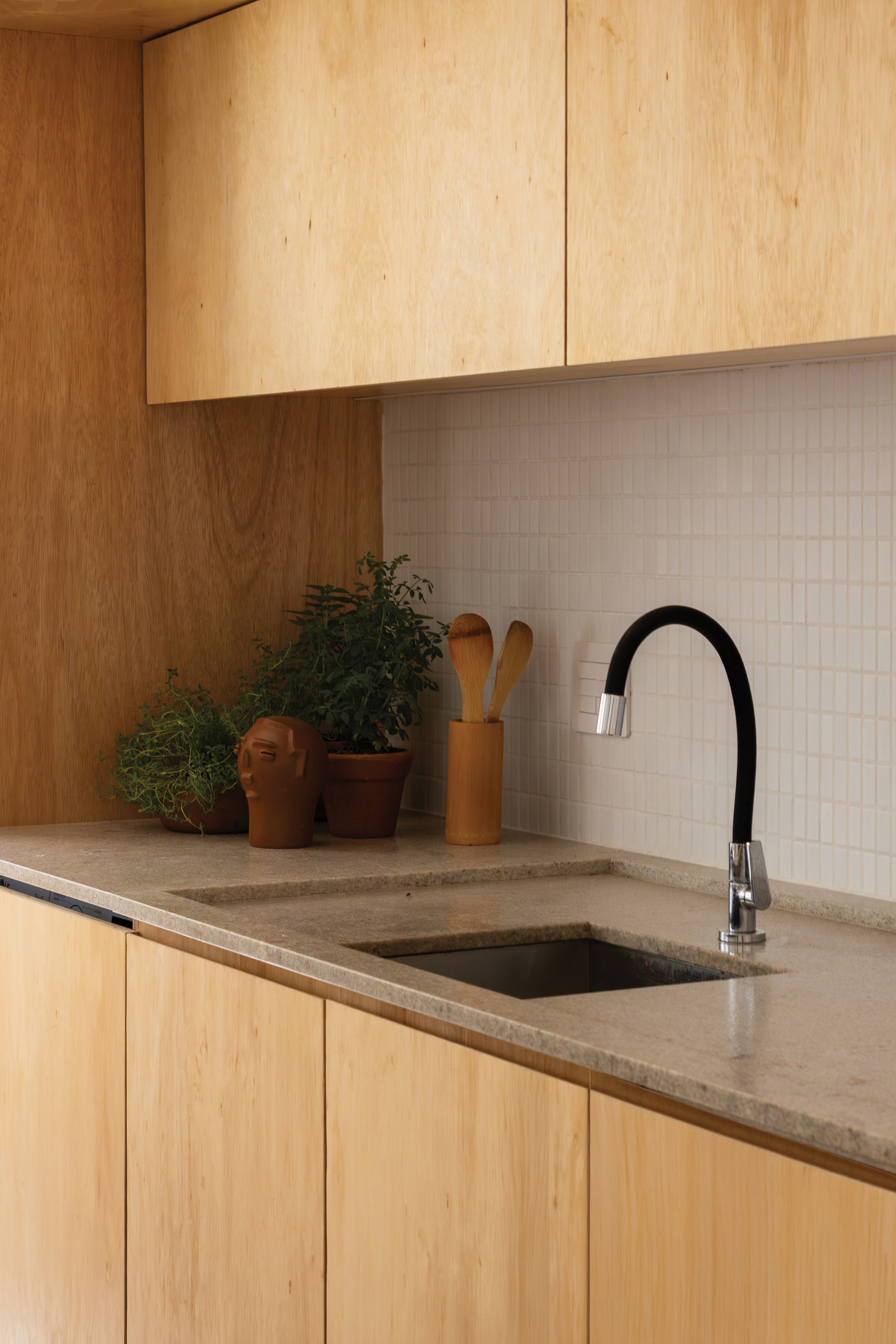 Close-up of the stone countertop, undermount sink and black gooseneck tap against white mosaic tile, with potted herbs
